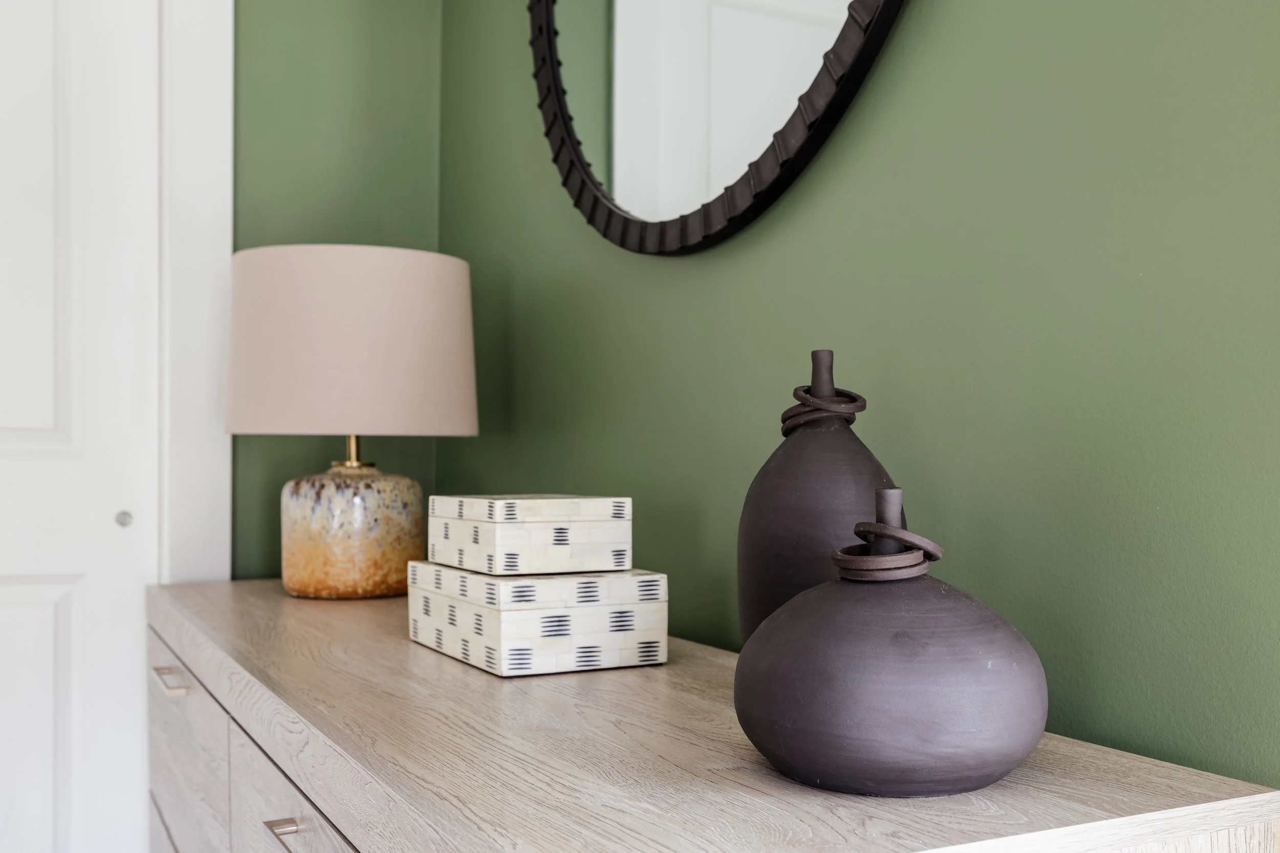 Decorative arrangement on a wooden dresser with two dark brown ceramic vases, a stack of patterned boxes, and a table lamp with a beige shade in front of a green wall and oval mirror.