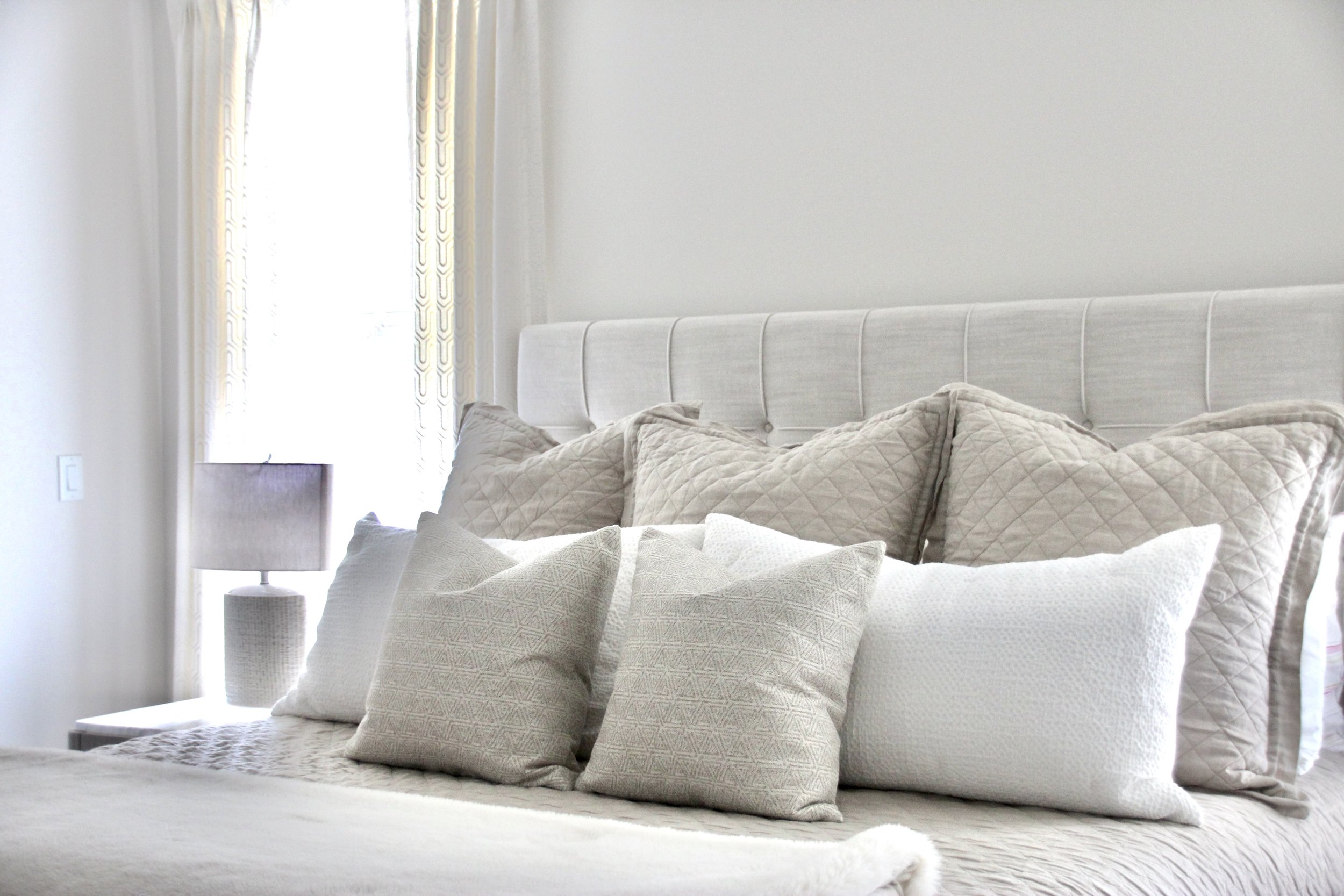 Modern bedroom interior with a light gray tufted headboard, several decorative pillows in shades of gray and white, a gray quilted bedspread, and a bedside lamp with a matching gray base.
