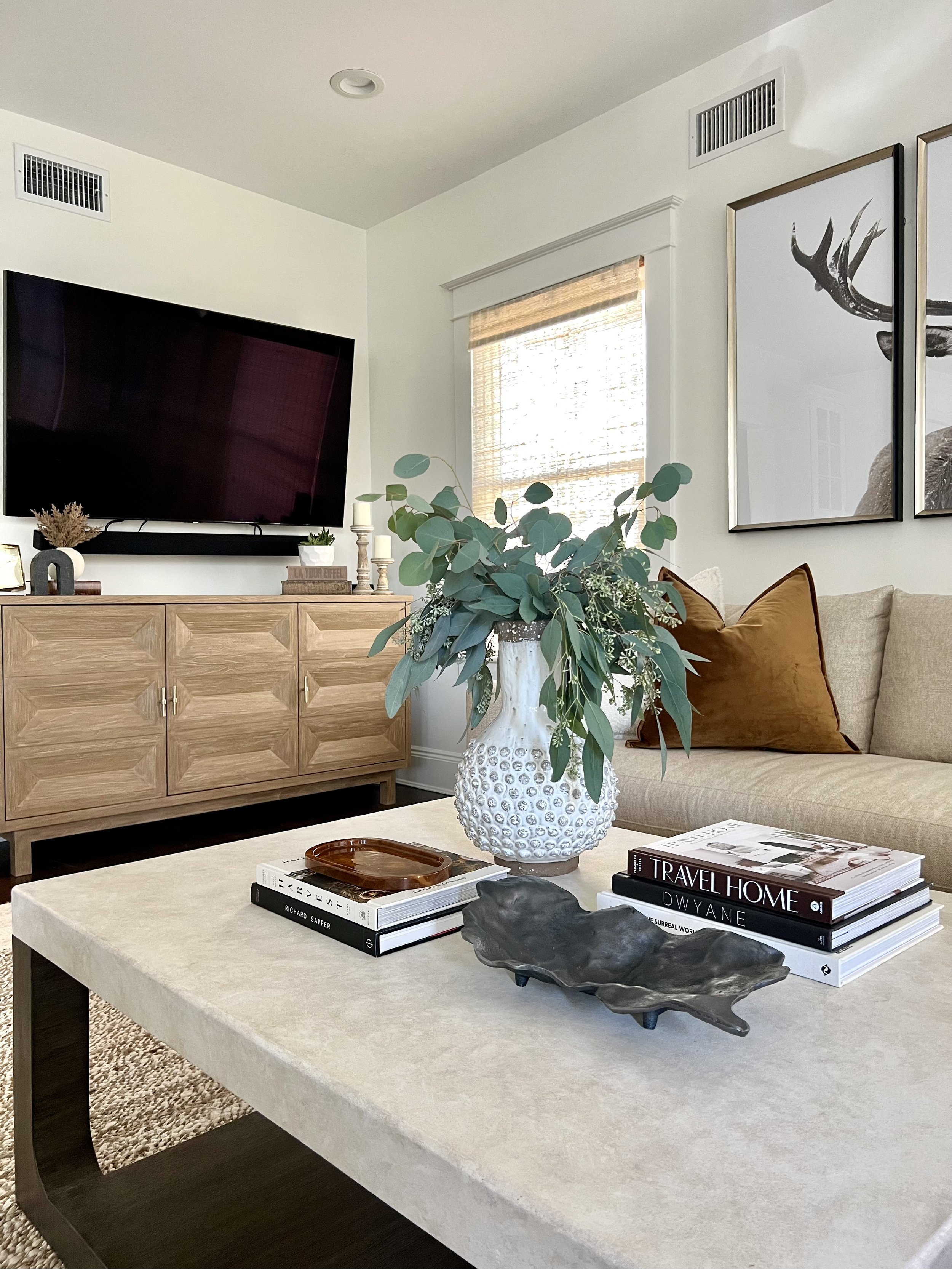 Modern living room with a mounted TV, wooden cabinet, gray sofa, decorative tray, books, and a vase with greenery on a coffee table.