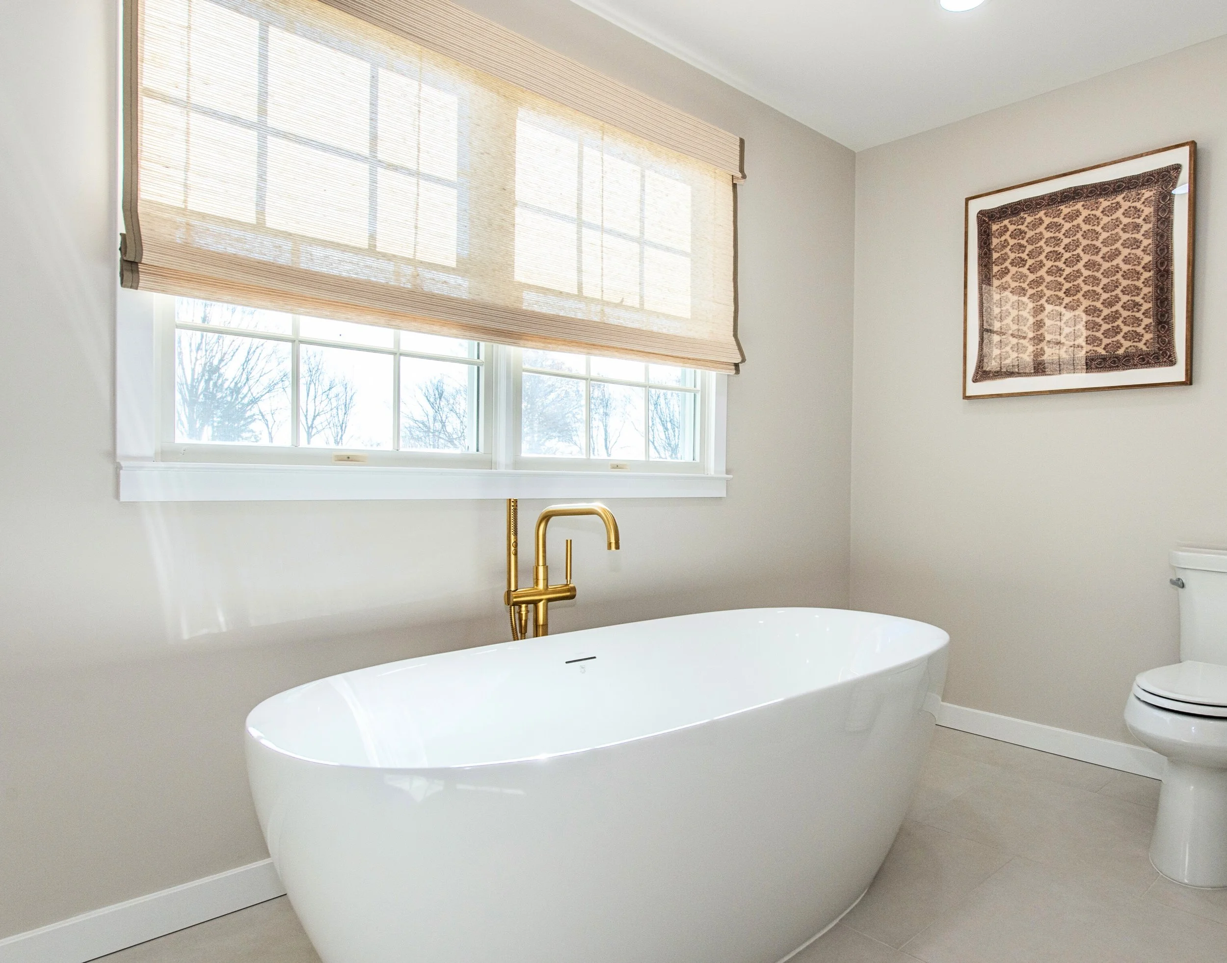 Modern bathroom with freestanding bathtub, gold faucet, window with beige shades, wall art, and toilet.