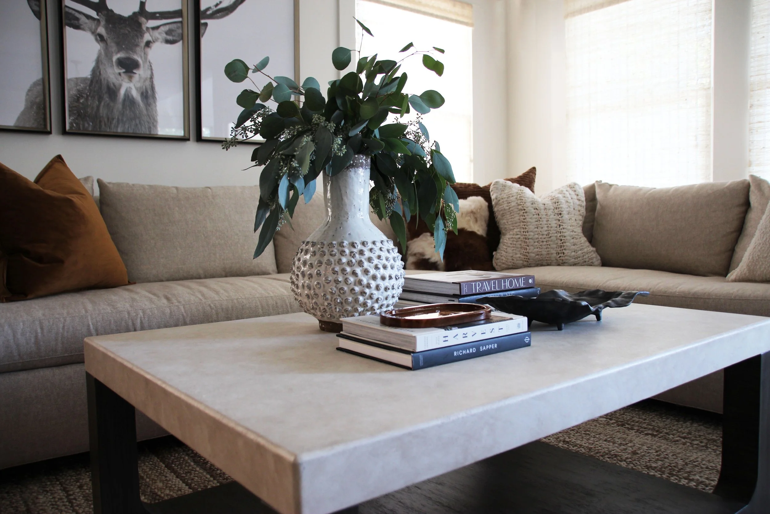 Modern living room with beige sectional sofa, decorative pillows, textured vase with greenery on coffee table, and art featuring deer on wall.