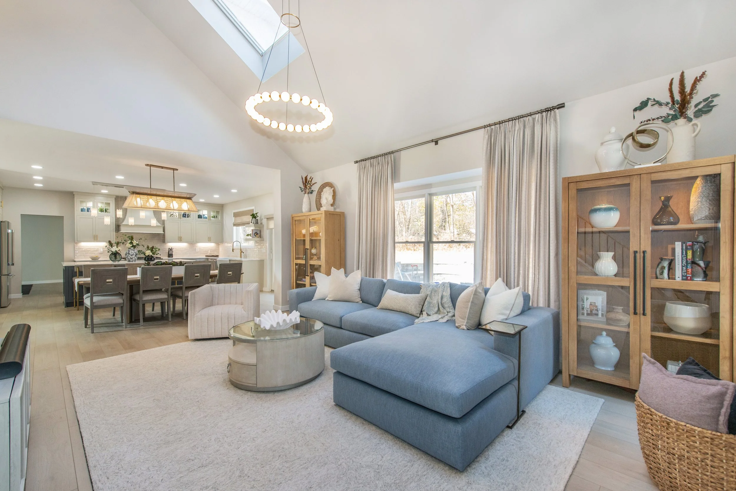 Modern living room with a blue sectional sofa, light-colored area rug, glass-top coffee table, and wooden display cabinet. Open-concept layout extends to a kitchen with an island and bar stools under modern lighting. Large window and curtains allow n