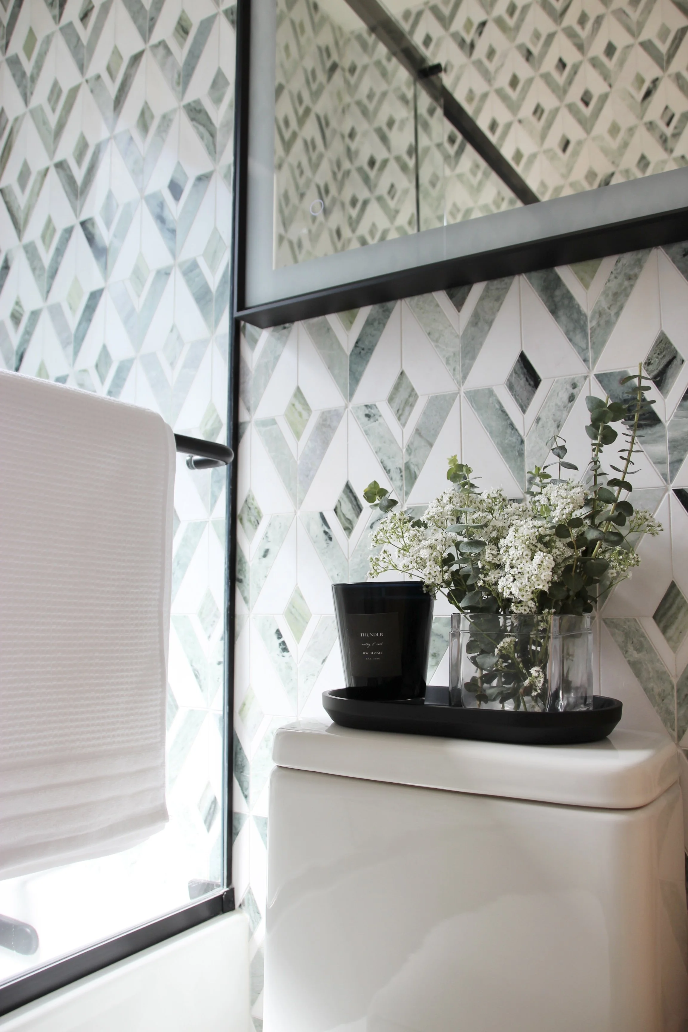 Modern bathroom with geometric tile wall featuring green and white patterns, a black-framed mirror, a white towel on a rail, and a black tray holding a black candle and a glass vase with white flowers and greenery on a toilet tank.