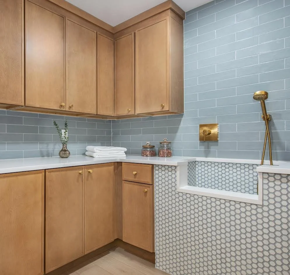 Modern laundry room with wooden cabinets, blue subway tile backsplash, white countertop, brass faucet, and tiled dog washing station.