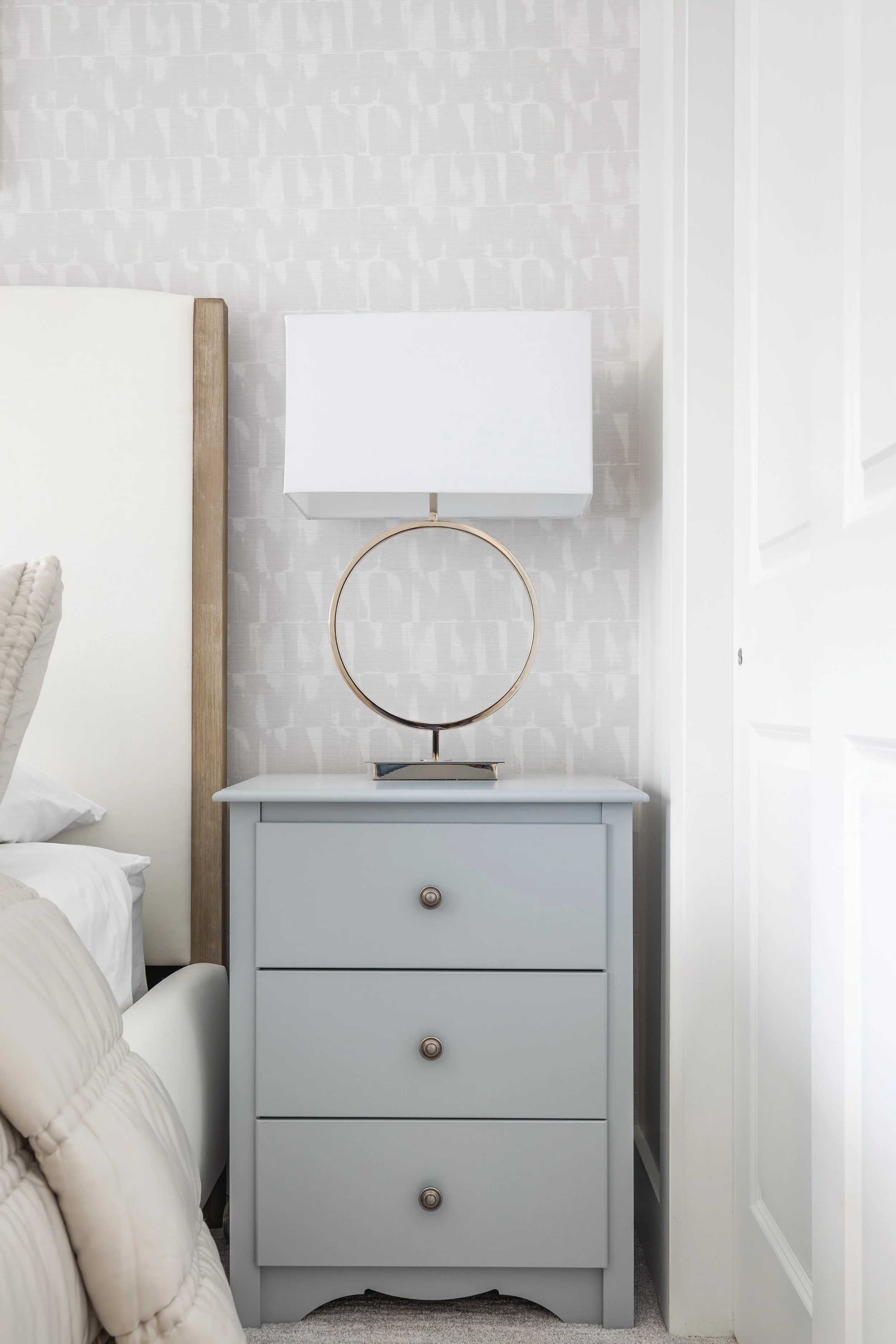 Modern bedroom with a light gray nightstand, three drawers, a circular gold lamp with a rectangular white shade, beside a bed with white bedding and a padded headboard, against a patterned wallpaper.
