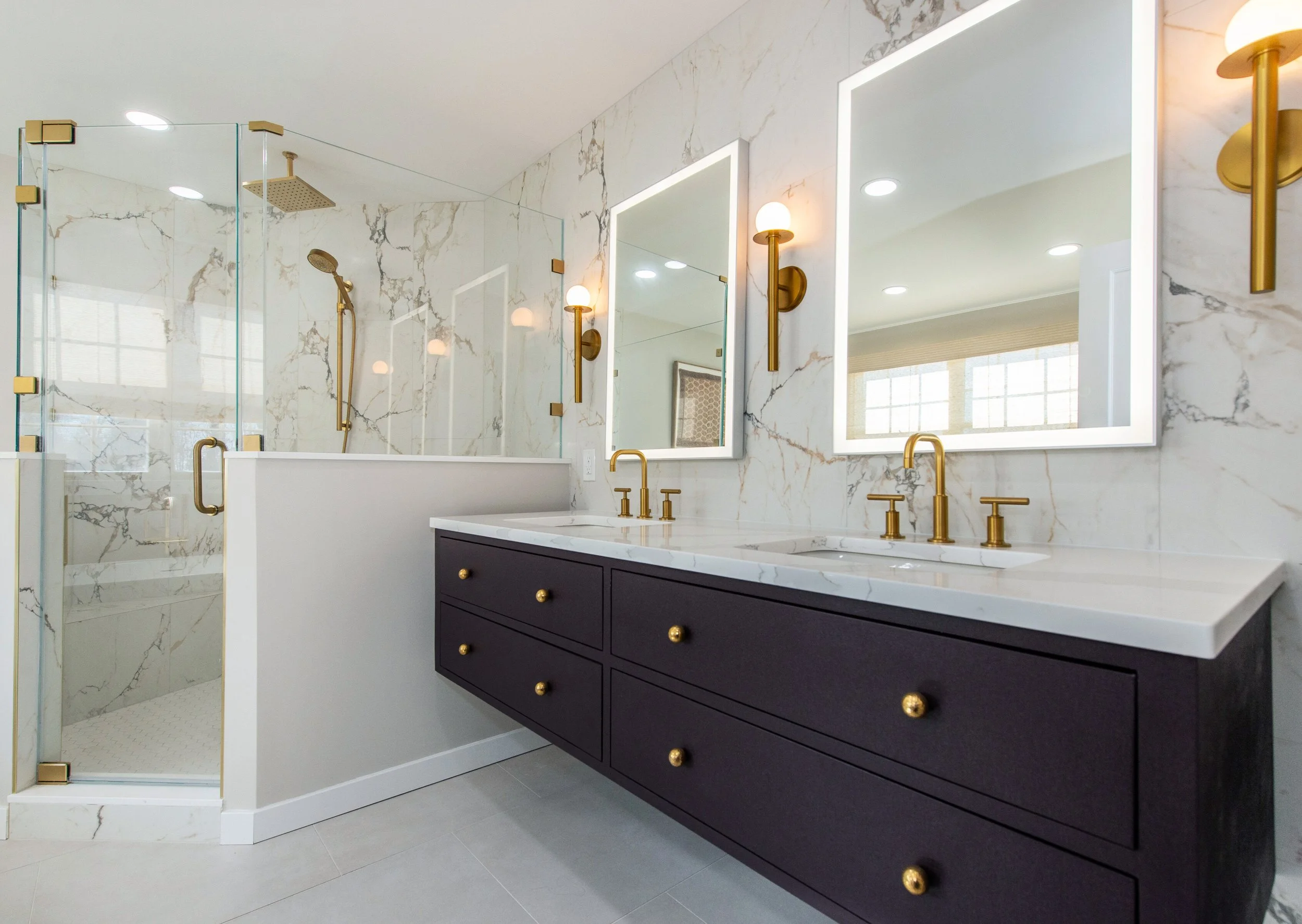 Modern bathroom with double sink vanity, gold fixtures, wall-mounted mirrors, marble tiles, and glass shower enclosure.