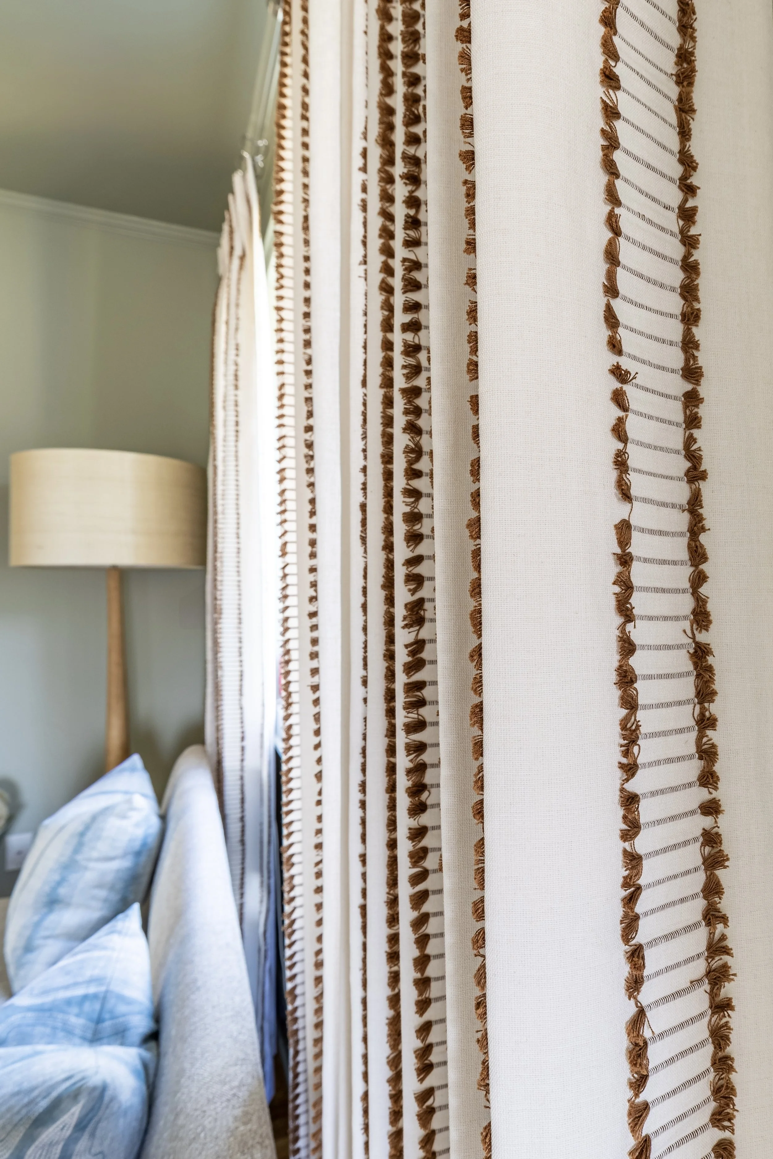 White curtains with brown tassel trim, beside a beige couch with blue pillows and a floor lamp.