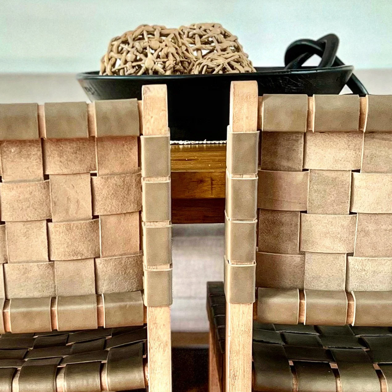 Close-up of two woven chairs made of leather straps and wood next to each other, with a black bowl containing woven decorative spheres in the background.