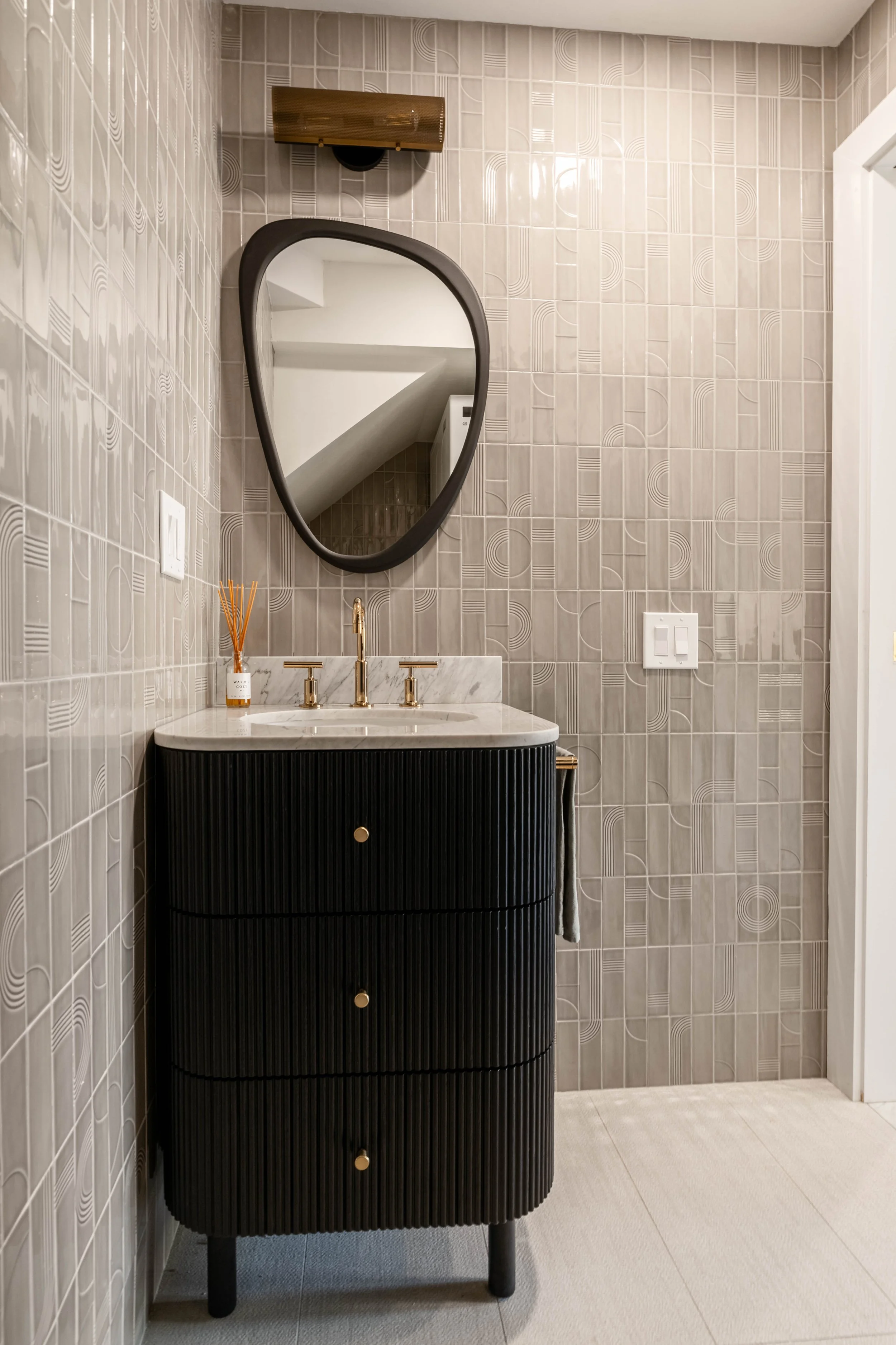 Modern bathroom interior with textured beige tile walls, a black ribbed vanity with a marble countertop, a gold faucet, and a geometric mirror above. Decorative bottle with reed diffuser on the counter and wall-mounted light fixture above the mirror.