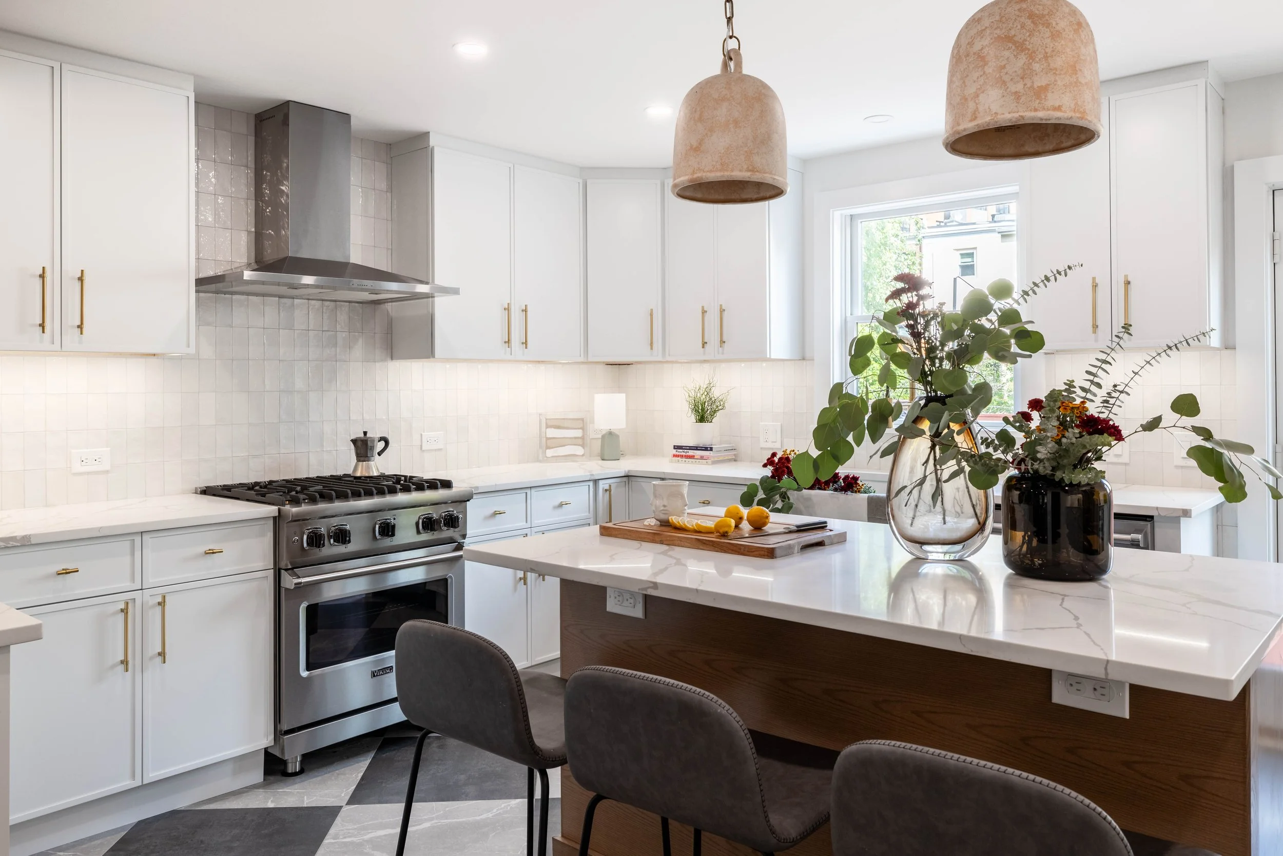 Modern kitchen with white cabinets, stainless steel stove, and two pendant lights over an island. The island has vases with flowers and a cutting board with lemons.