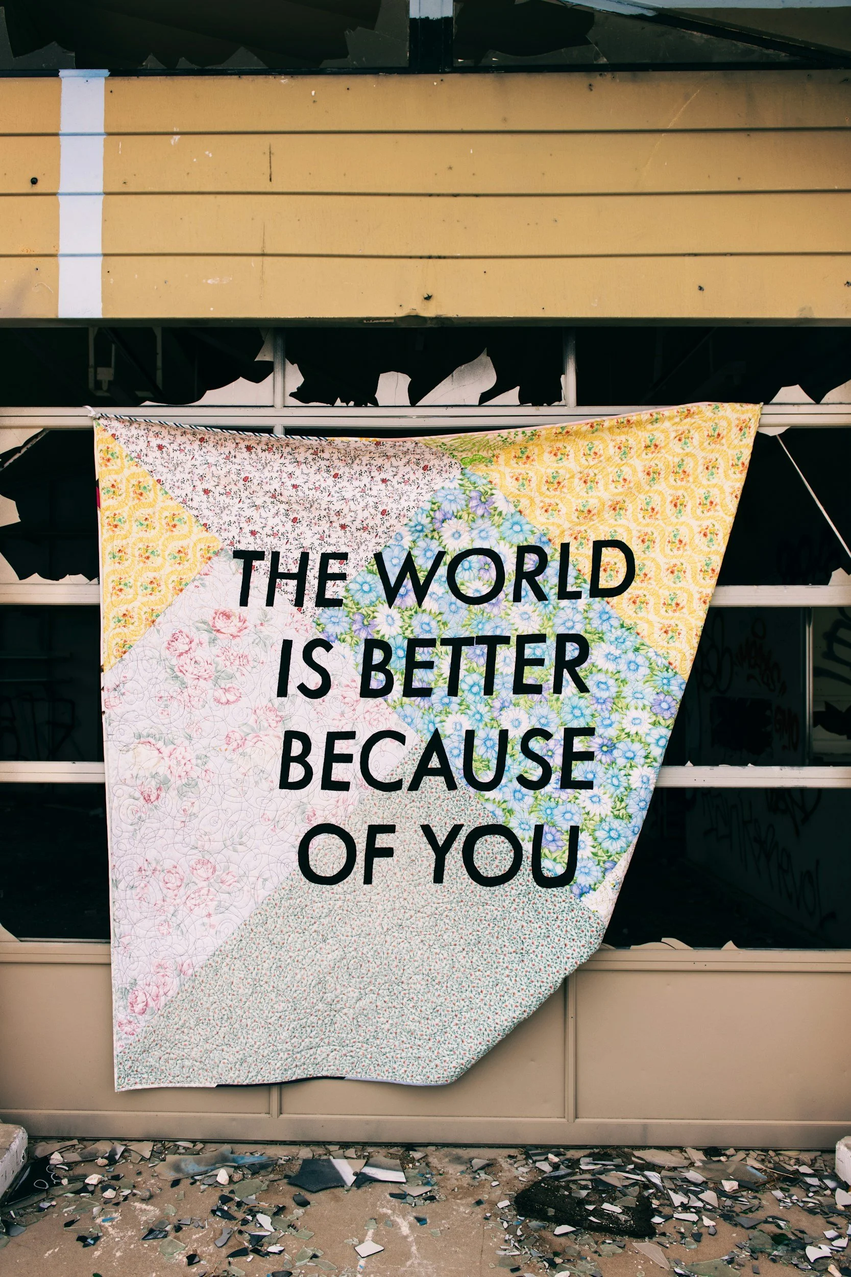 A quilt hanging on a broken, graffiti-covered structure with the message "The world is better because of you" stitched in black letters.