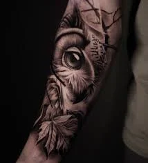 Black and gray tattoo of an owl with detailed feathers and large eyes, surrounded by leaves, on a person's arm.