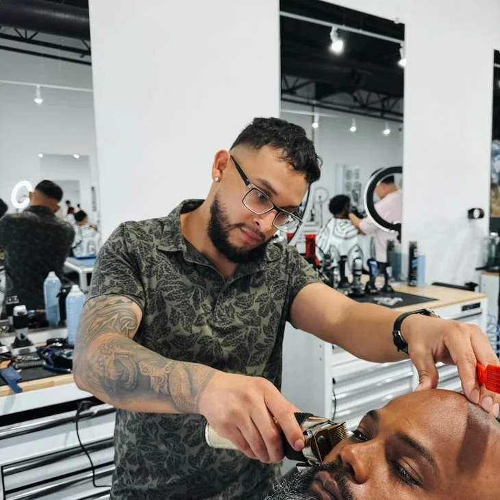 Barber cutting hair of a client in a modern barbershop with other patrons and mirrors in the background.