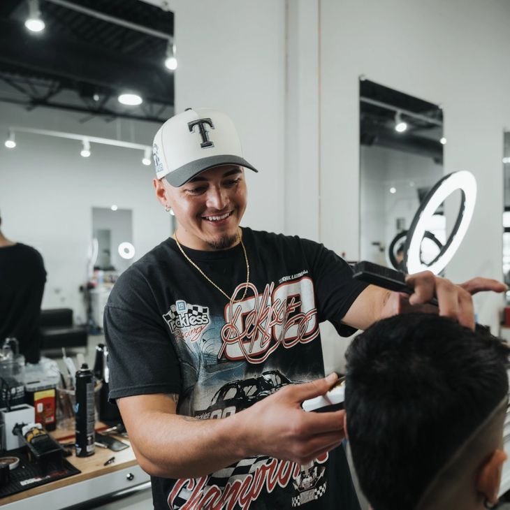 A smiling barber or hairstylist in a black graphic t-shirt and white baseball cap giving a haircut to a customer in a modern salon with a mirror, ring light, and hair products on the counter.