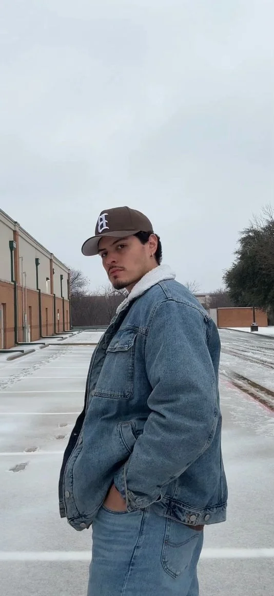 A young man wearing a baseball cap, denim jacket, and jeans standing in an empty parking lot on a cloudy winter day.