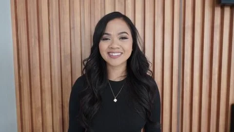 A woman with long dark hair, wearing a black top and a necklace, smiling in front of a wooden wall.