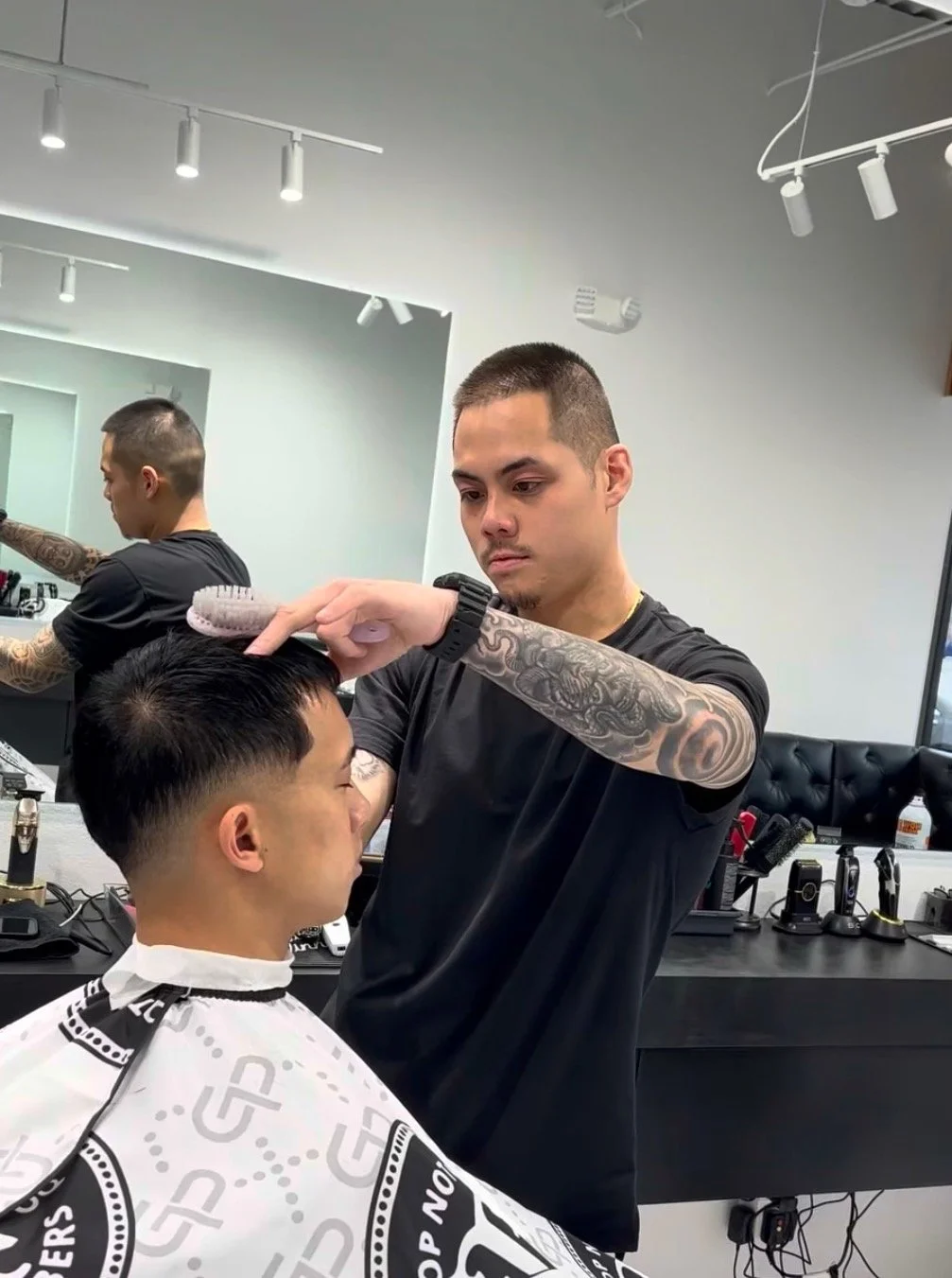 Barber giving a haircut to a young man in a salon, with hairdresser using a brush and mirror in the background.
