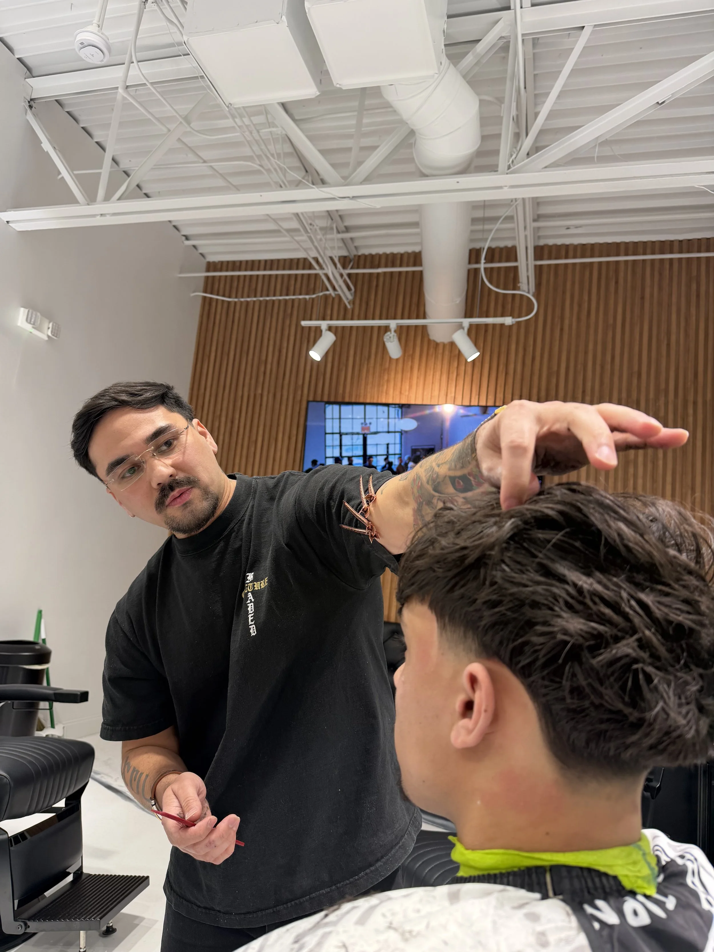 Barber giving a haircut to a young man in a modern barber shop with wooden wall paneling and exposed ceiling pipes.