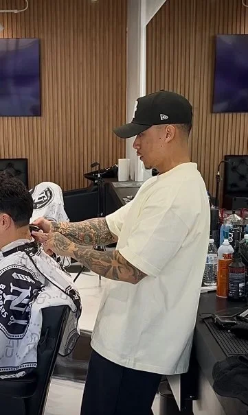 A man with tattoos on his arms wearing a black baseball cap and white t-shirt getting a haircut in a barbershop.