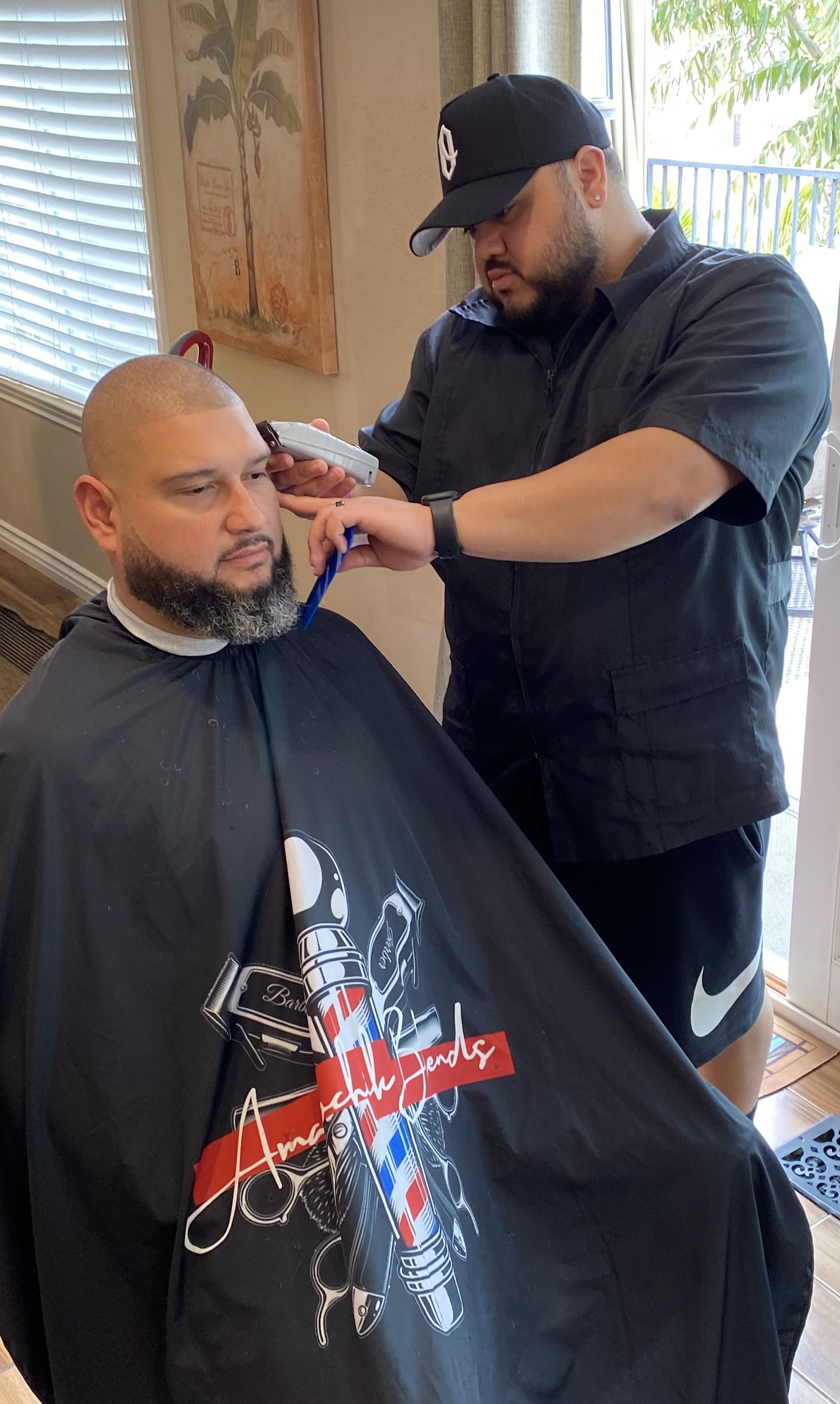 Man receiving a fade haircut from a barber in a barbershop, seated with a black barber cape on. The barber is focused, wearing a cap, black shirt, and shorts, using clippers on the man's hair. The barbershop has a window with blinds, artwork on the wall, and natural light coming in.