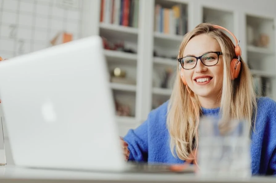 A woman wearing glasses and pink headphones working on a laptop in a bright, organized room with bookshelves in the background.