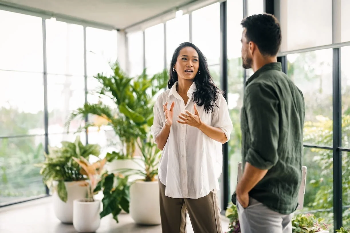 A woman and a man having a conversation indoors with large windows and potted plants in the background.