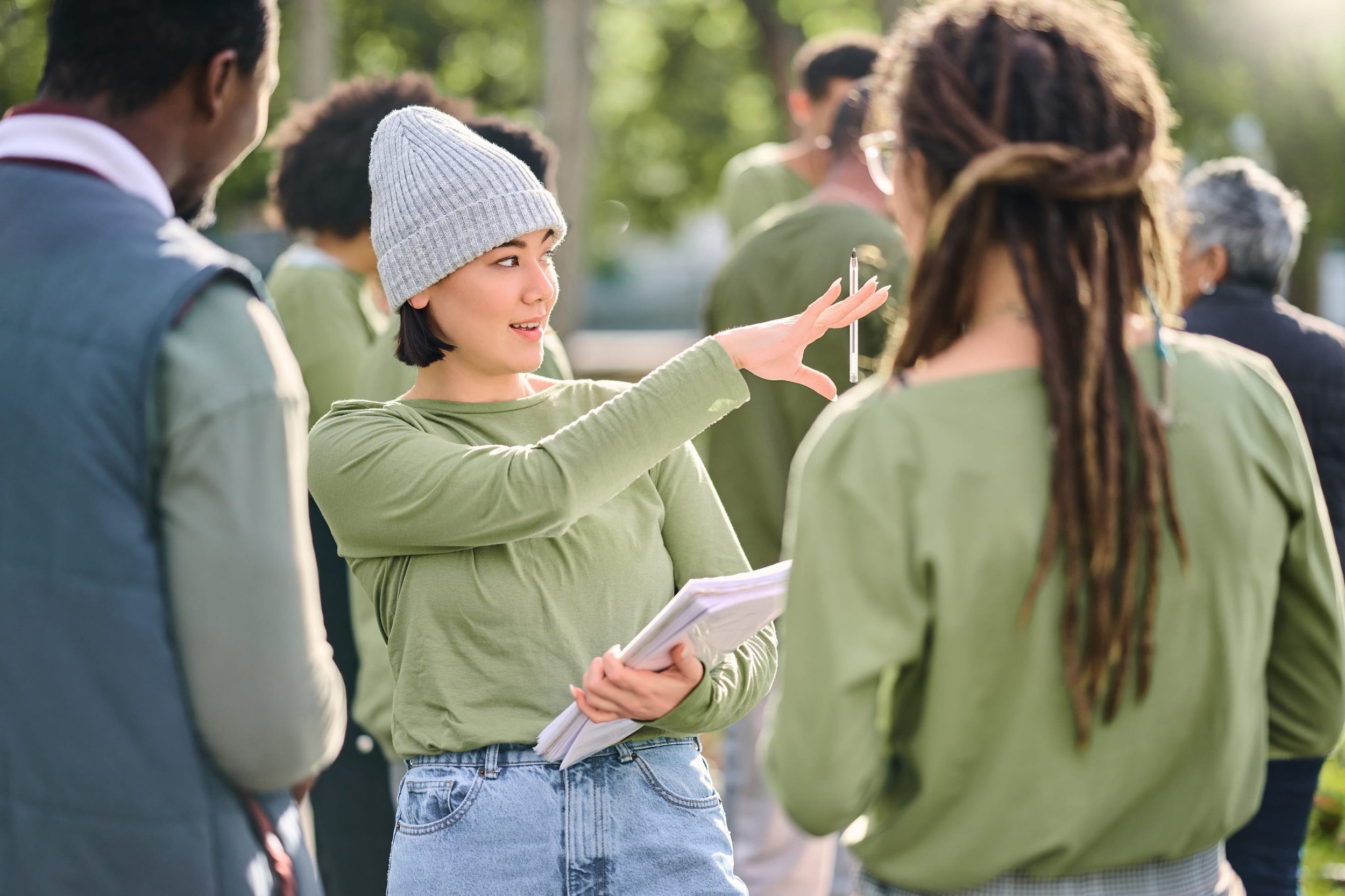 A young woman in a gray beanie and green shirt holding papers and a pen, speaking to a group of people outdoors.