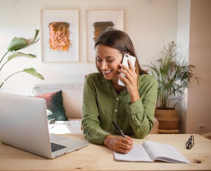 A woman sitting at a desk, talking on the phone, writing in a notebook, with a laptop open in front of her, in a cozy home office with plants and abstract art on the wall.