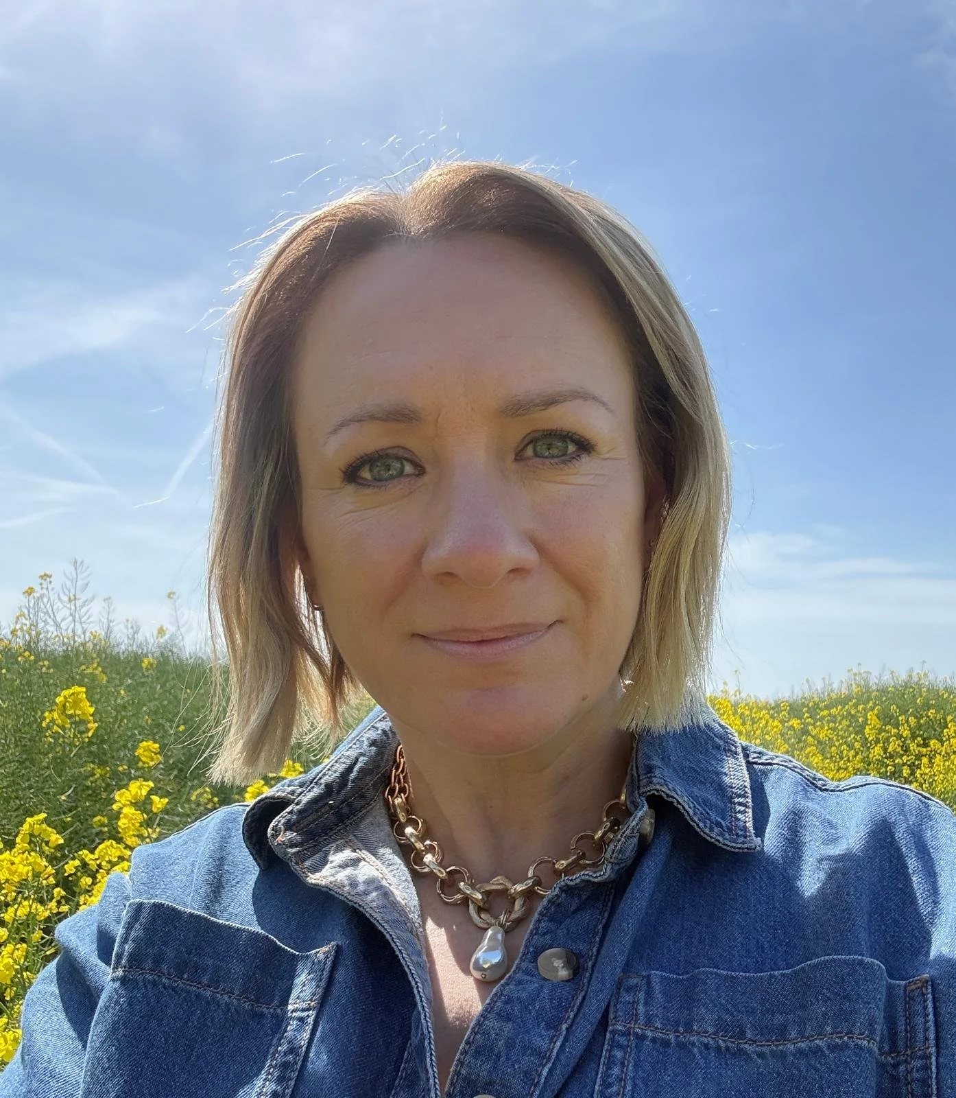 Dawn Foster, founder of We Do Wellbeing, outdoors in a field of yellow flowers with a blue sky and clouds behind her.