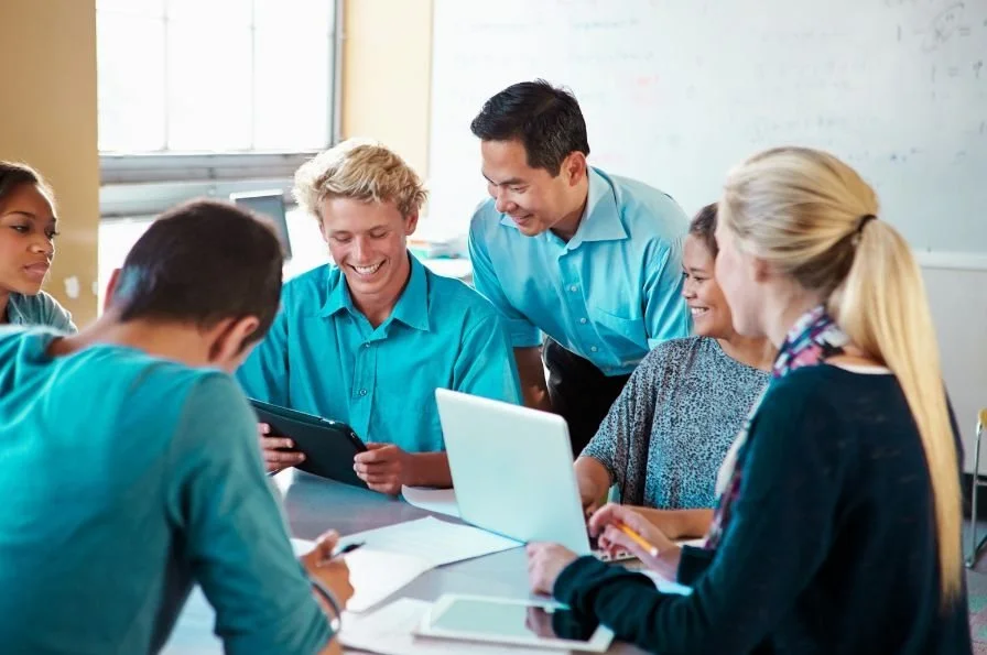 A diverse group of students and a teacher gathered around a table in a classroom, looking at a tablet and a laptop.