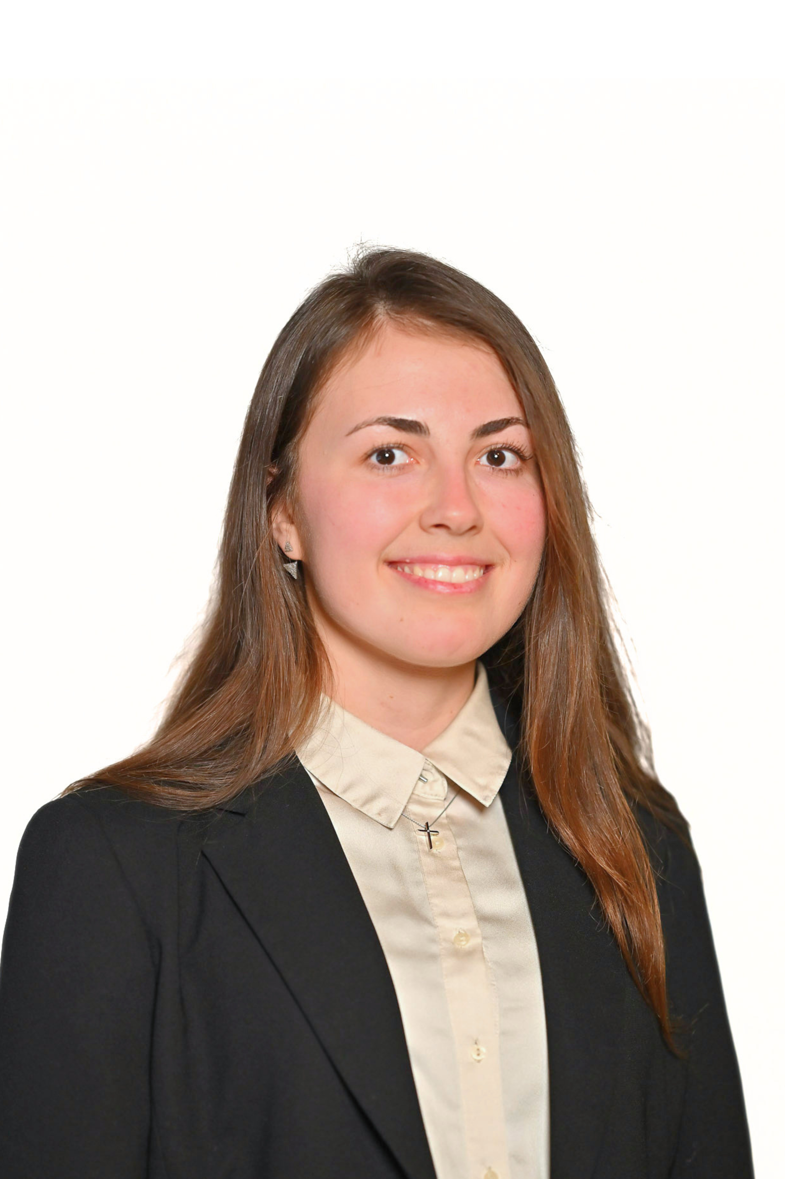 A woman with long brown hair wearing a black blazer and a light-colored shirt, smiling in front of a plain background.