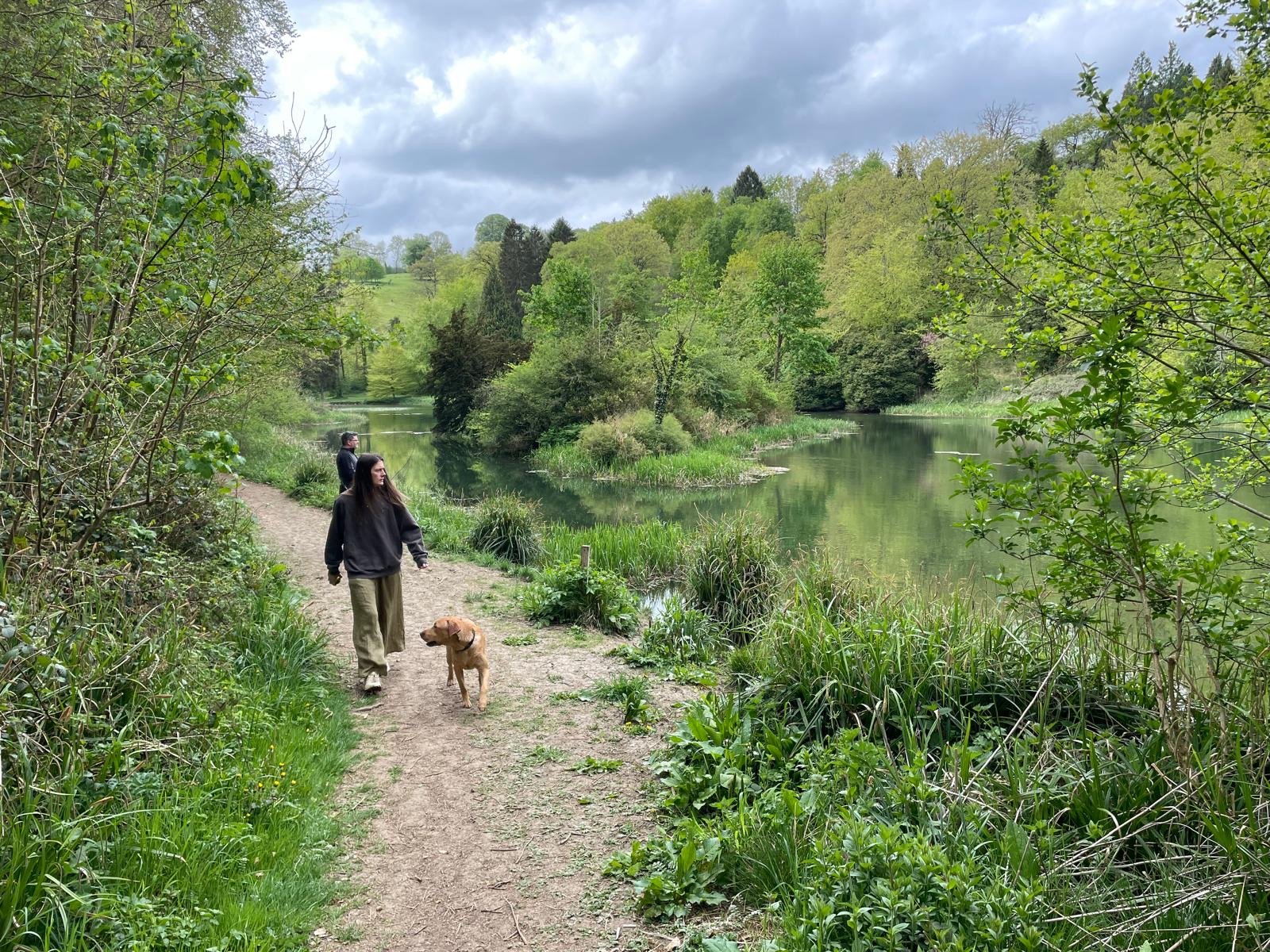 Two women and a dog walking along a dirt trail beside a calm river in a lush green forest under a cloudy sky.