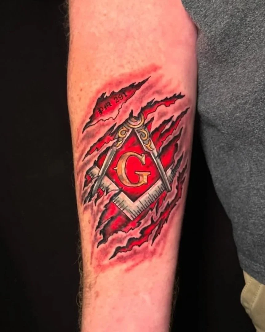 A tattoo on a person's forearm featuring a Masonic symbol with a red and black background, a ruler and compass intertwined, and the letter G in the center. There are fiery red and black accents around the symbol.