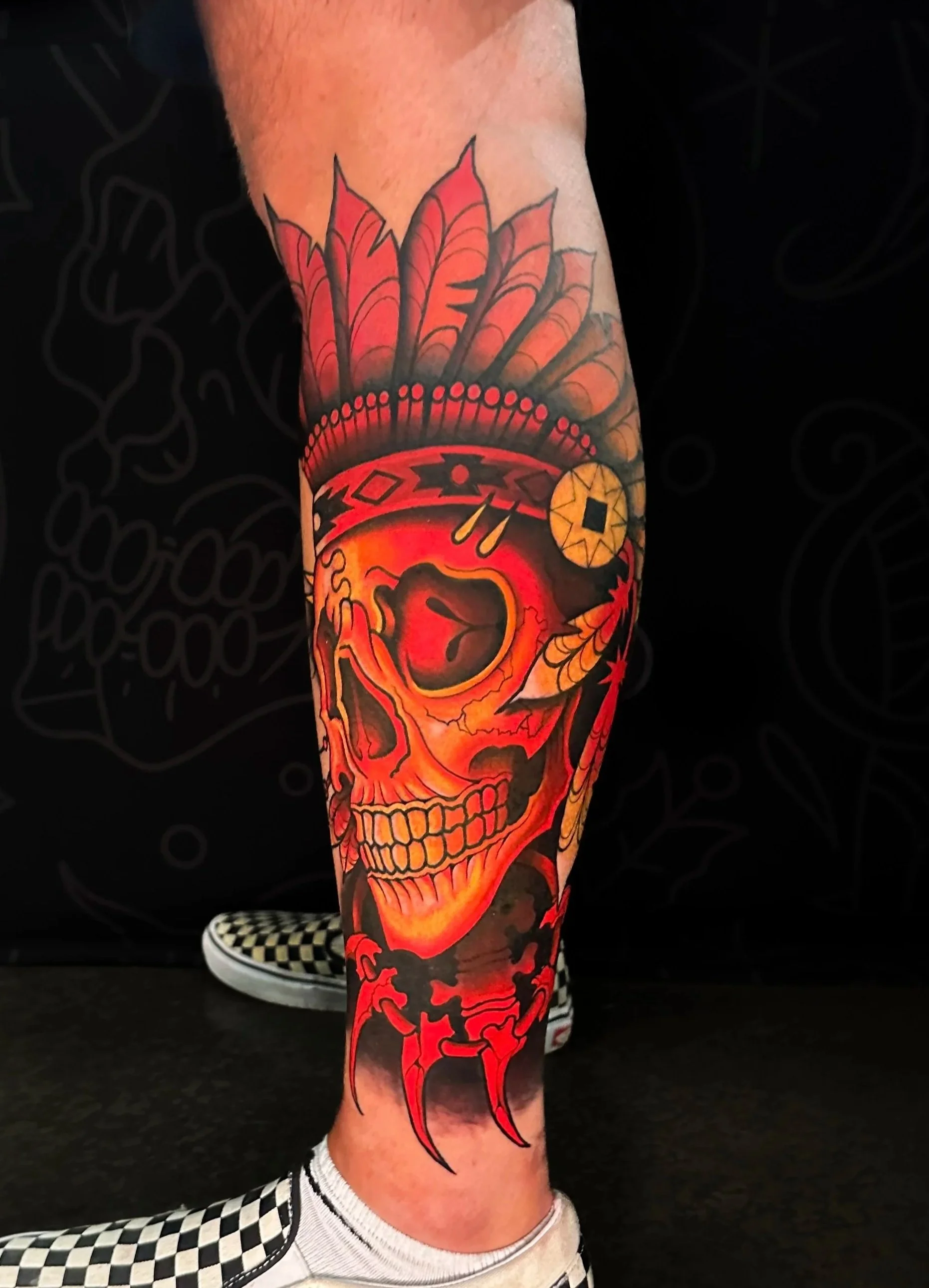 Colorful tattoo of a skull with a flaming glow on its face, wearing a feathered headdress, and a bone necklace. on a person's lower leg.