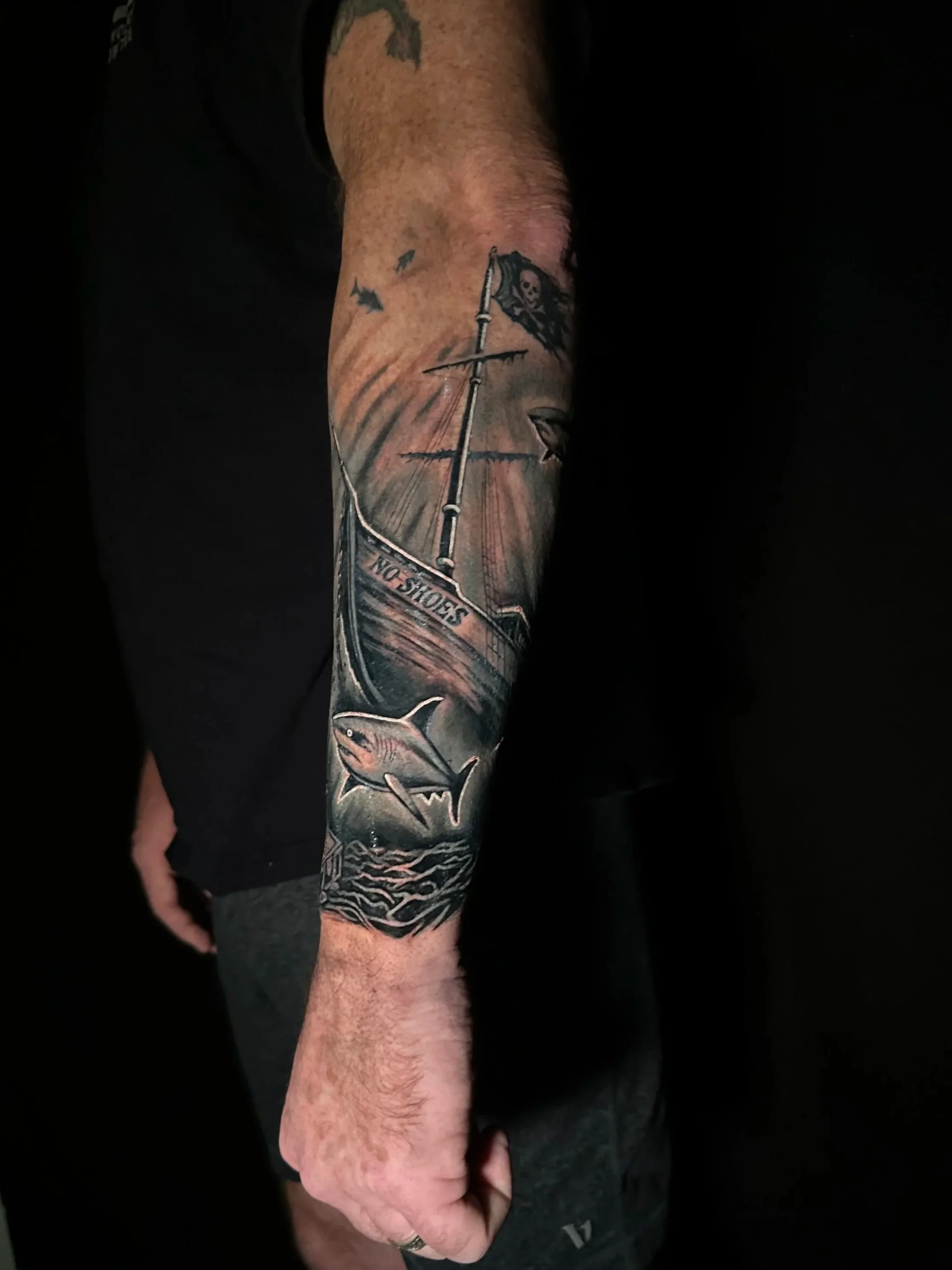 Tattoo of a sailing ship with a skull flag, a shark, and the words 'NO SHOES' on the person's forearm.