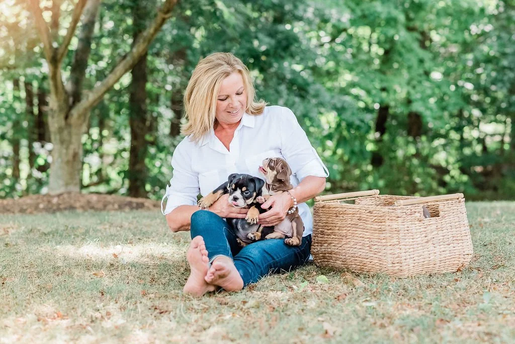 A woman sitting on the grass outdoors playing with two puppies, with a basket next to her and lush green trees in the background.