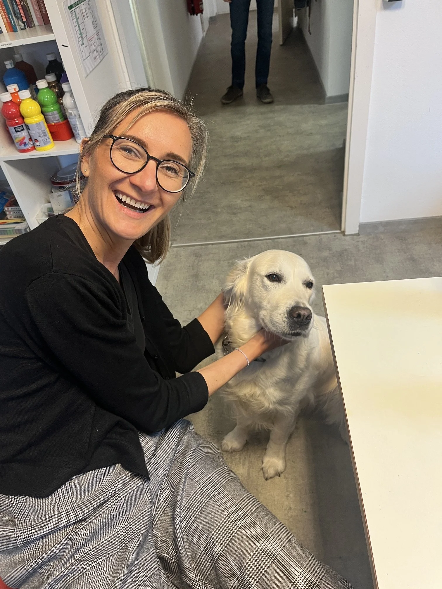 Eine lächelnde Frau mit Brille sitzt auf dem Boden in einem Innenraum und hält einen hellen Hund im Arm. Im Hintergrund sind ein Flur, ein Spiegel und ein Regal mit bunten Flaschen zu sehen. Die Stimmung wirkt warm und nahbar.