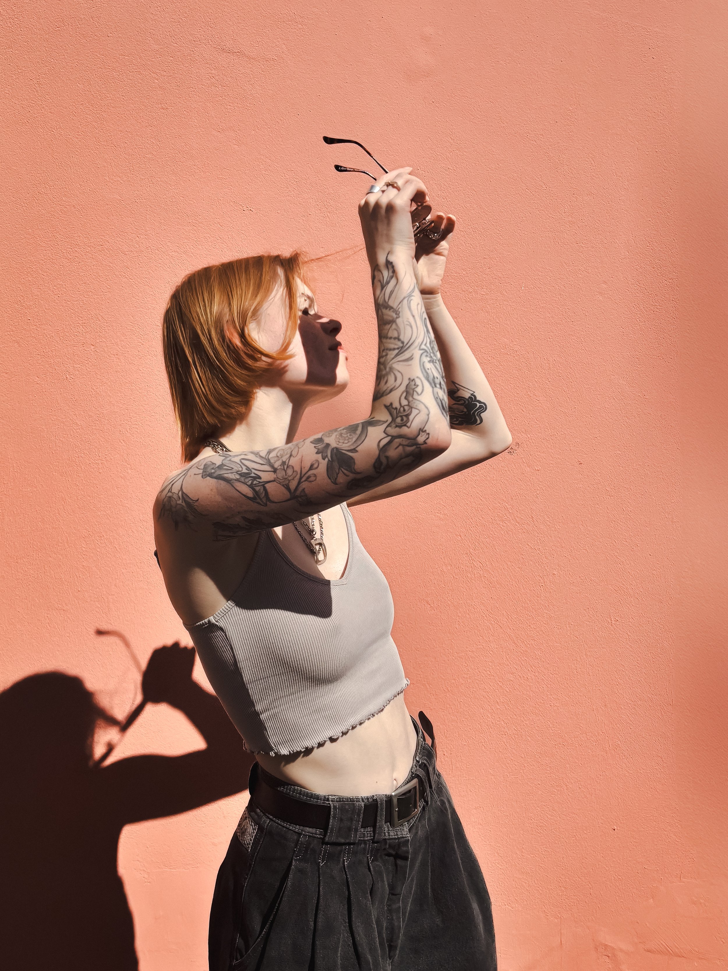 Eine Frau mit kurzen roten Haaren steht seitlich vor einer rosafarbenen Wand. Sie trägt ein graues Crop-Top und dunkle Hose, hat tätowierte Arme und hebt eine Sonnenbrille in Richtung Licht. Die Sonne wirft einen starken Schatten an die Wand.