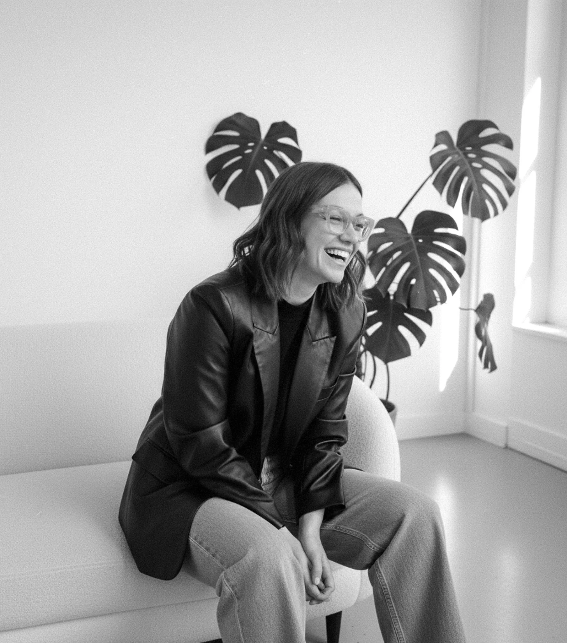Frau mit Brille lacht herzlich in schwarzem Lederblazer, sitzt mit Monstera-Pflanzen im Studio, Schwarz-Weiß-Foto.