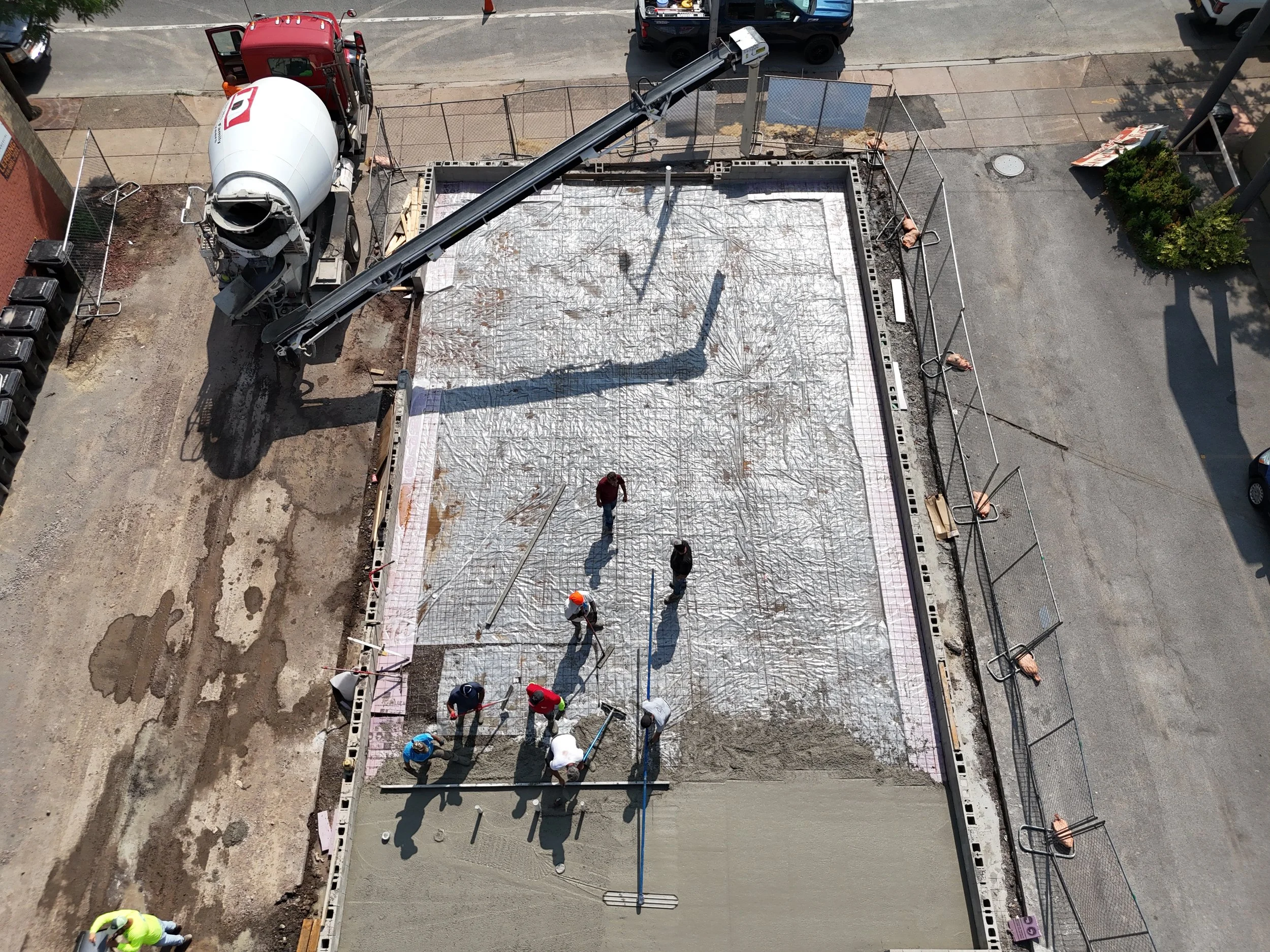 Construction workers pouring and leveling concrete on a building foundation, with a concrete mixer truck and a concrete pump nearby, and construction barriers surrounding the site.