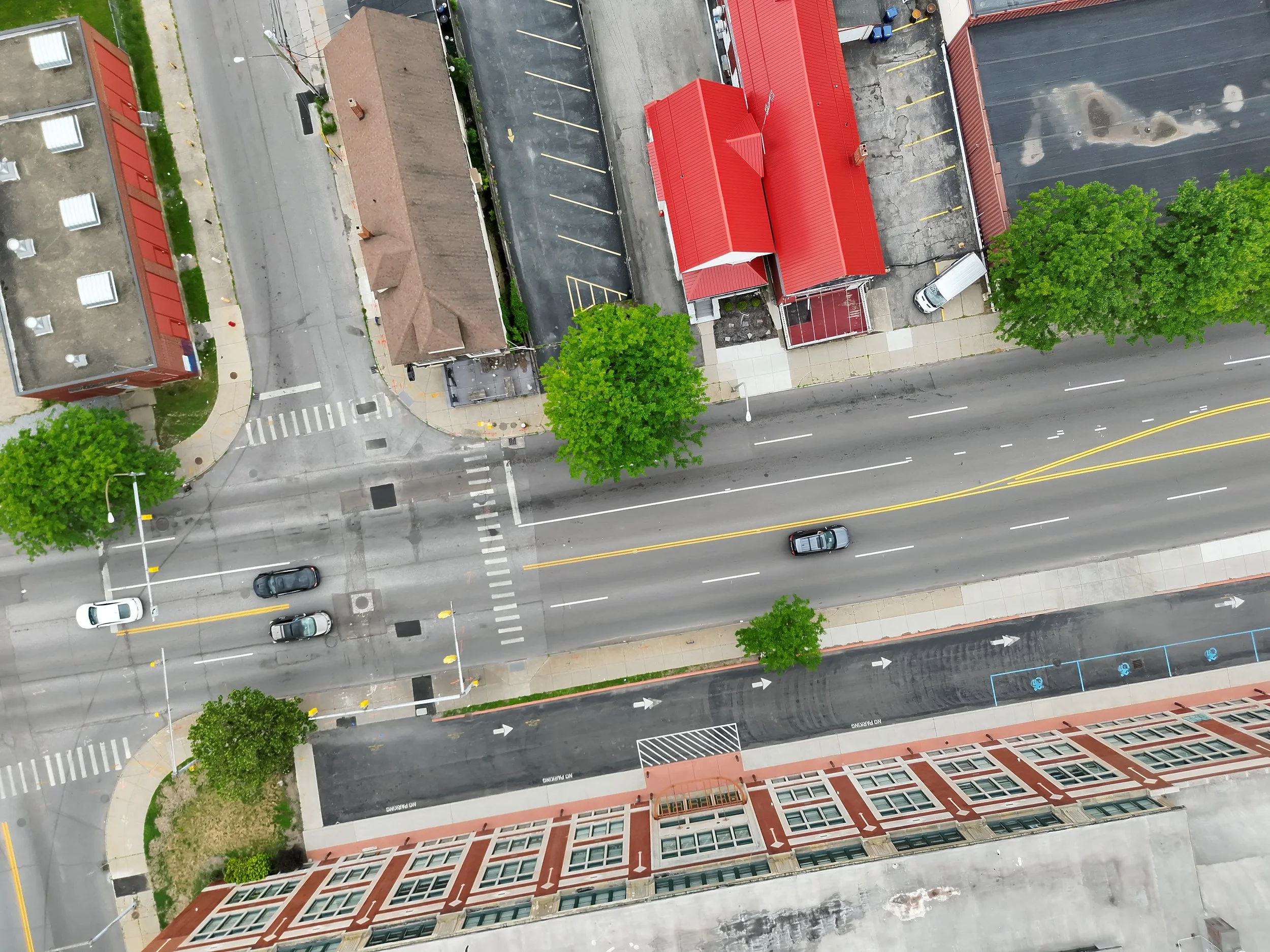 An aerial view of a city street intersection with a few cars, trees, buildings, and a parking lot with a red-roofed structure.