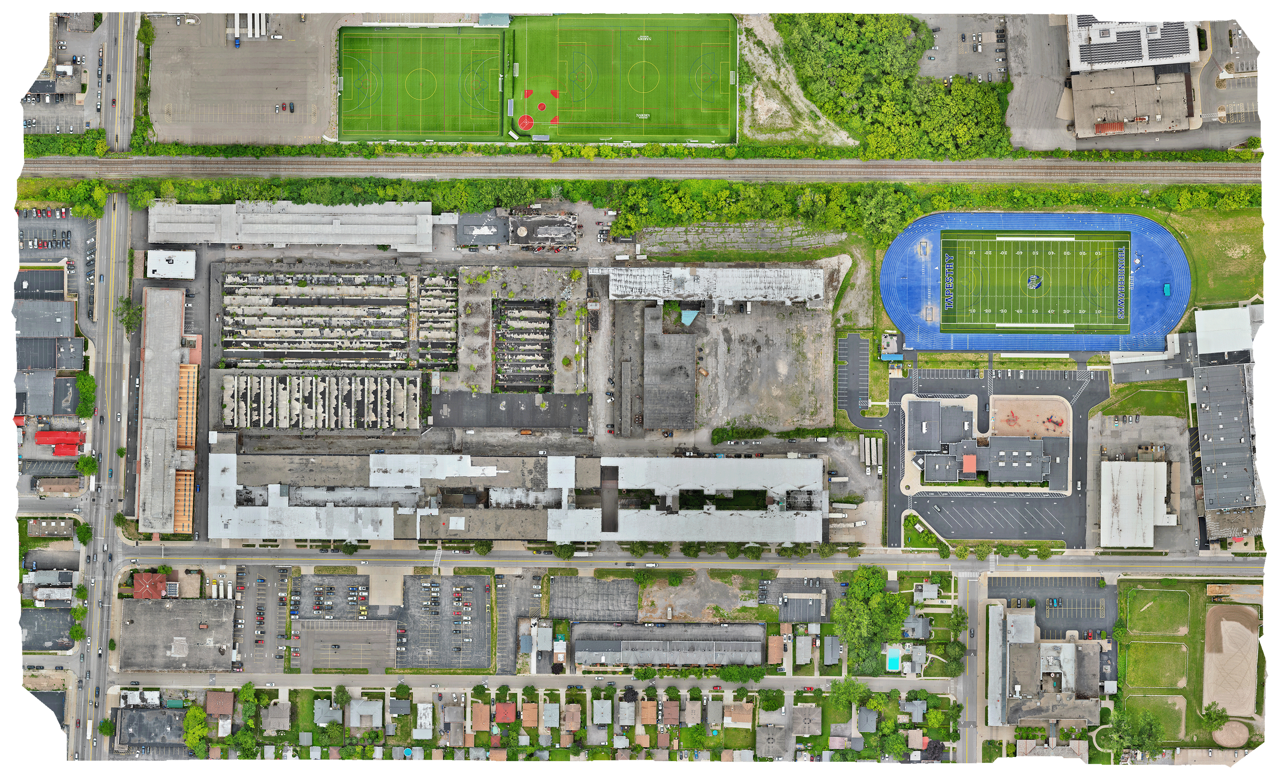 Aerial view of an urban area with sports fields, buildings, parking lots, and residential houses. Two sports fields are visible: one with a green soccer field and another with a blue track and football field. A cemetery with numerous graves is also visible. Streets with cars are on the left and bottom edges.