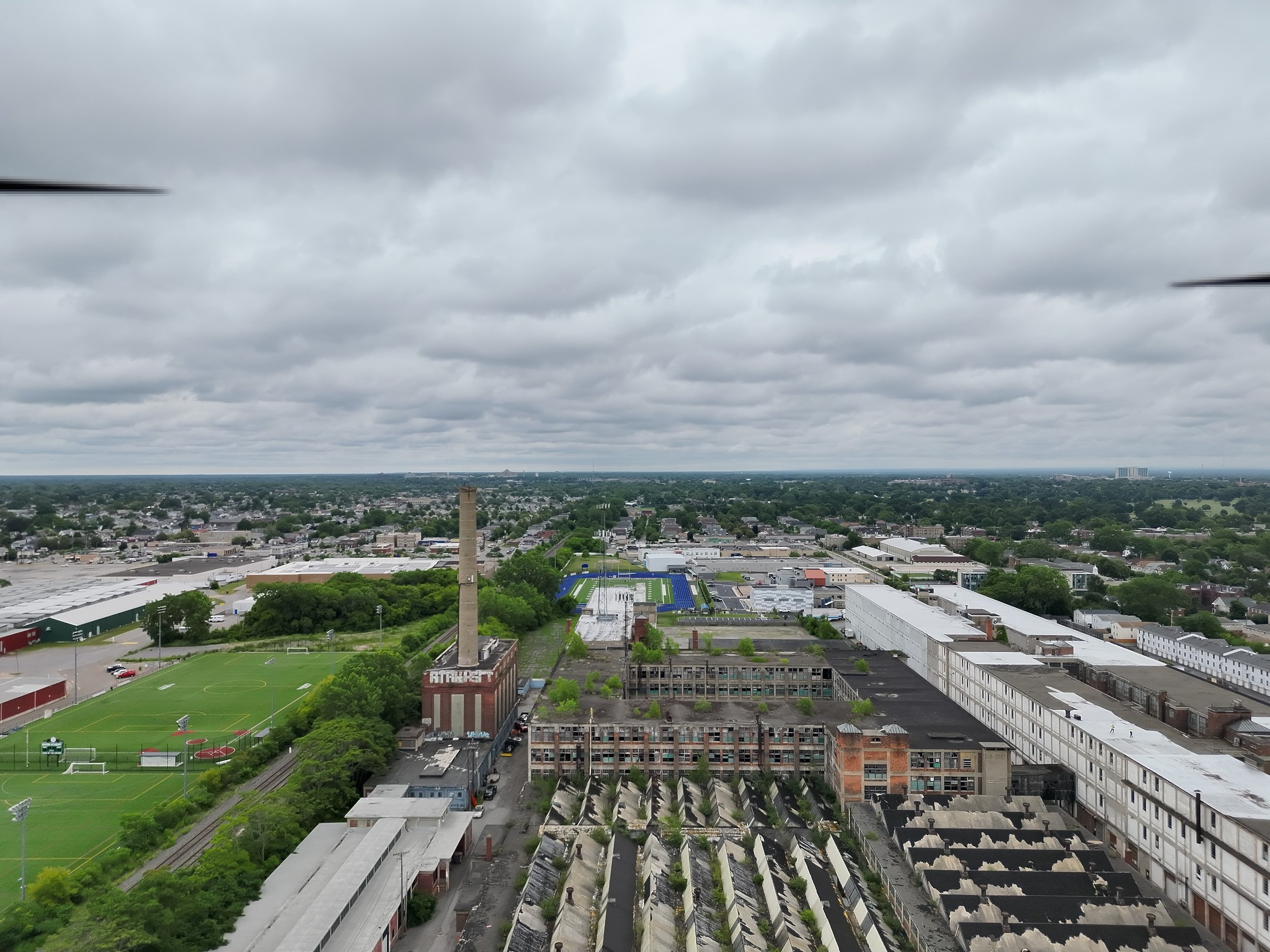 Aerial view of a cityscape on a cloudy day showing industrial buildings, a football field, and residential neighborhoods in the distance.