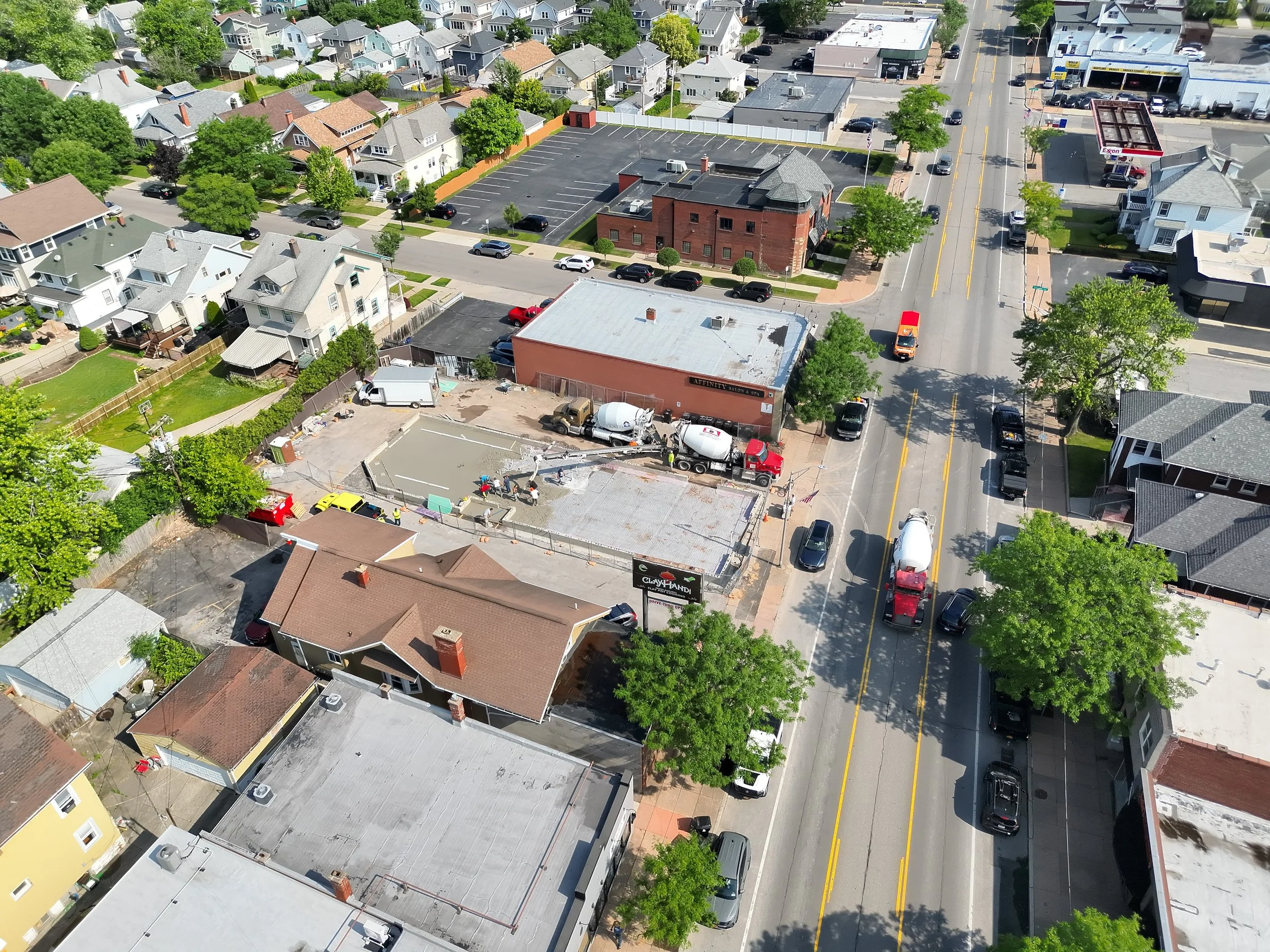 Aerial view of a small town street with commercial and residential buildings, trees, parking lots, and vehicles, including cement mixers and construction equipment, indicating ongoing construction.