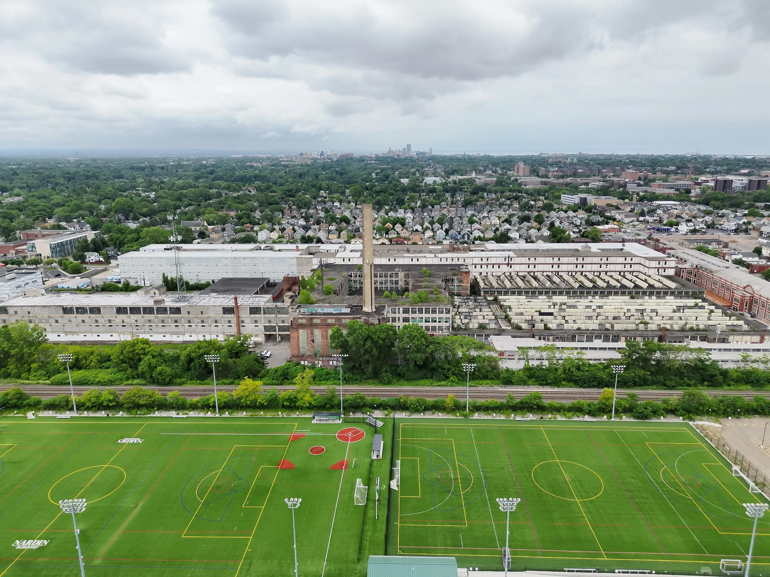 Aerial view of a sports complex with multiple soccer and football fields with vibrant green grass, surrounded by trees and city buildings with overcast sky.