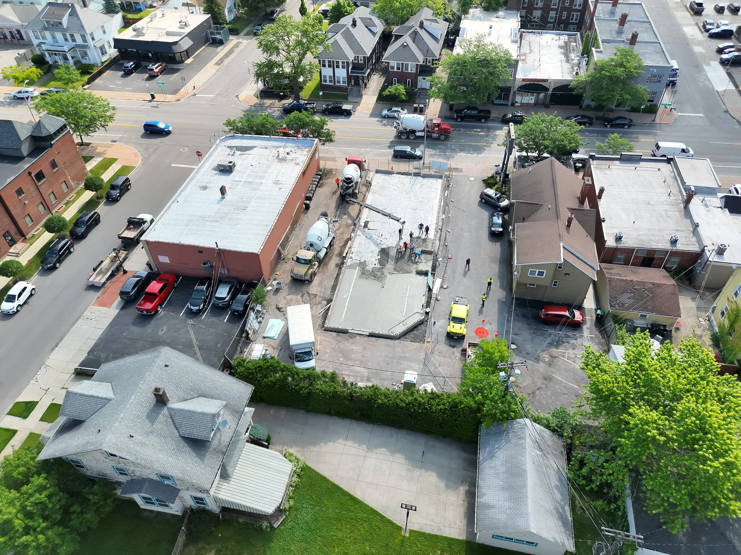 An aerial view of a construction site in an urban neighborhood, with workers and construction vehicles, surrounded by residential and commercial buildings, trees, and parked cars.