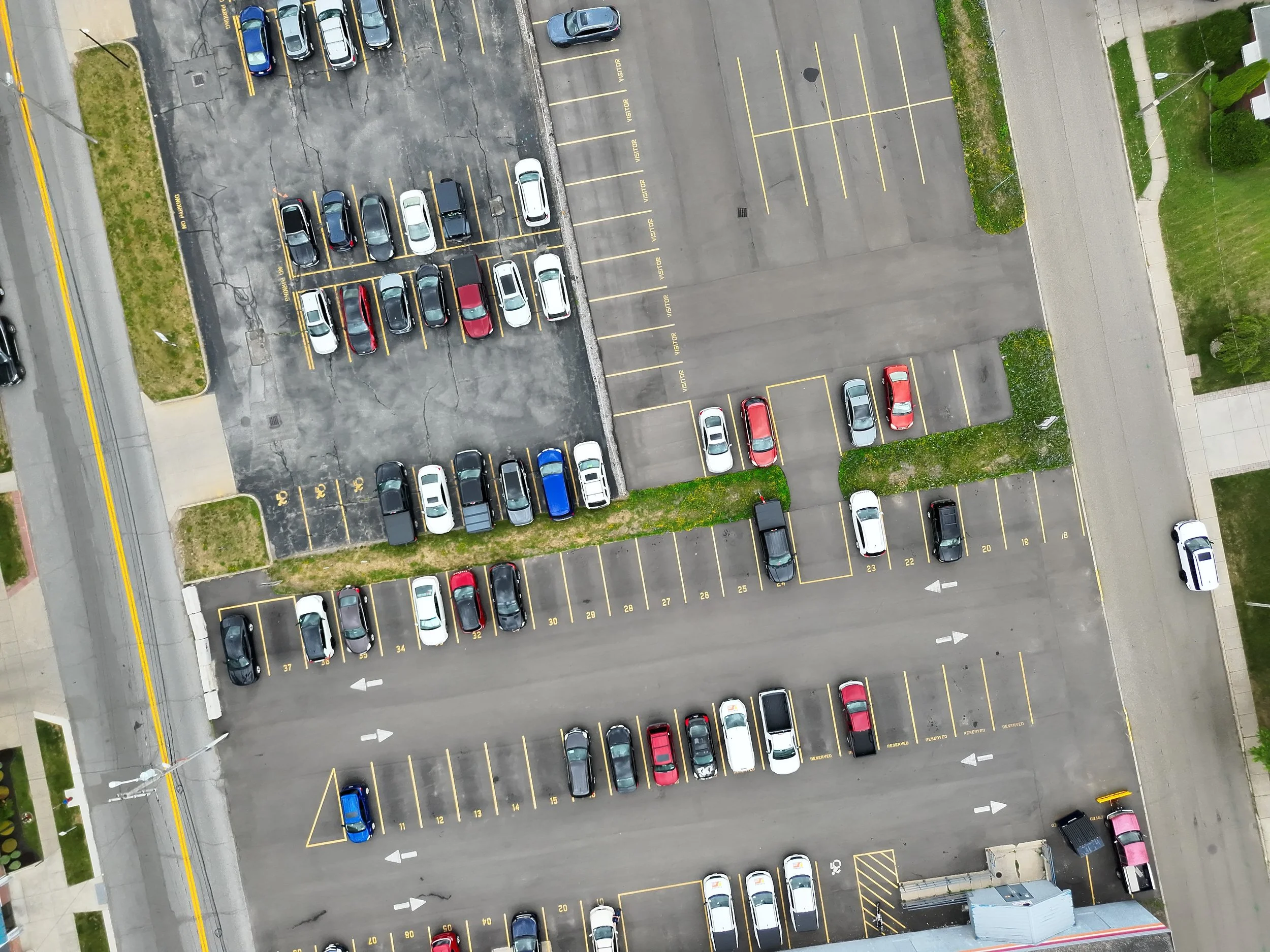 An aerial view of a parking lot with various cars parked in designated spaces, some labeled and others not, with surrounding sidewalks and grass areas.