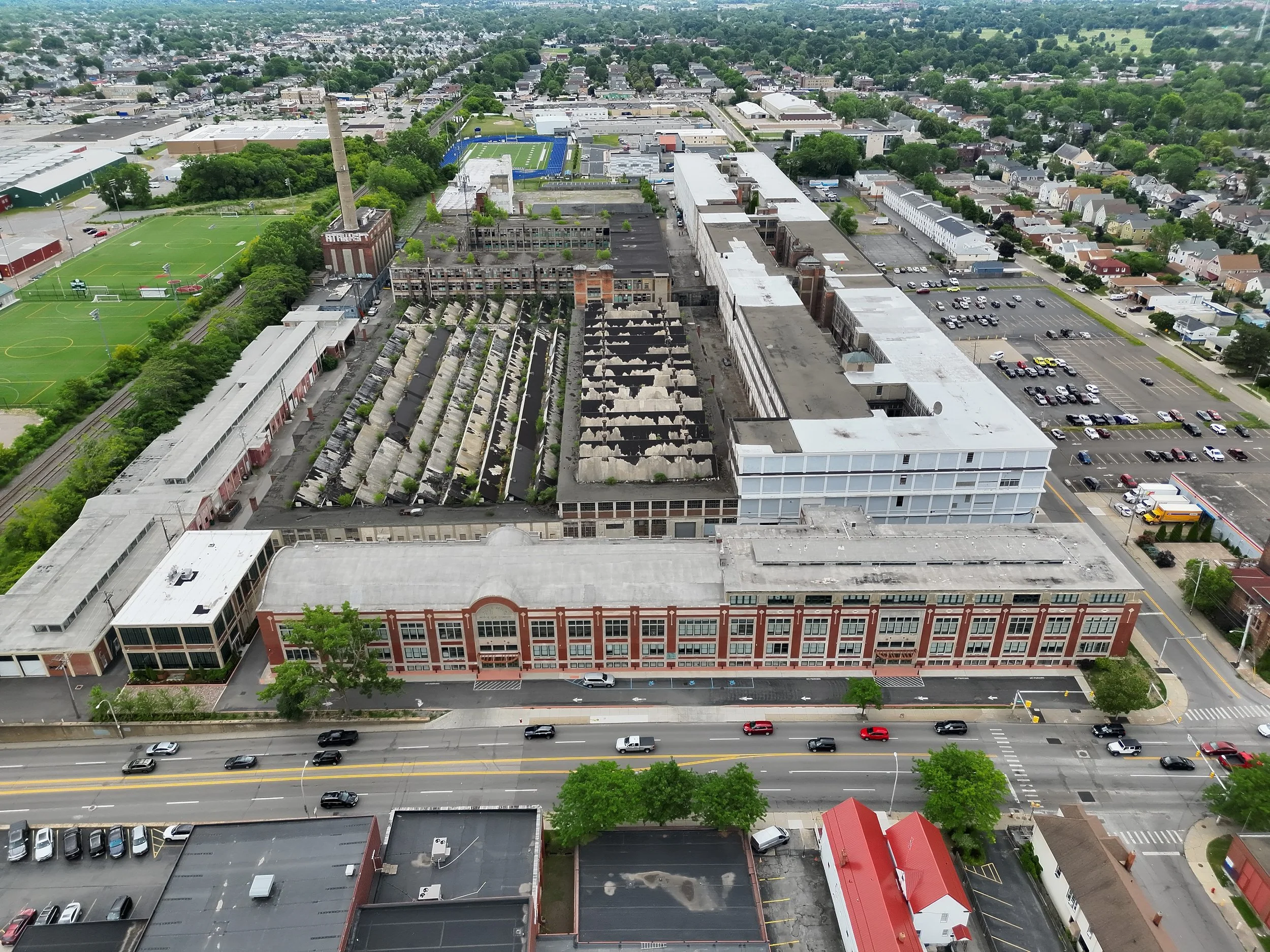 An aerial view of a cityscape showing a main street with cars, older brick buildings, a parking lot, another commercial building, an empty roof, and a sports field with a football field in a park area, surrounded by residential neighborhoods.