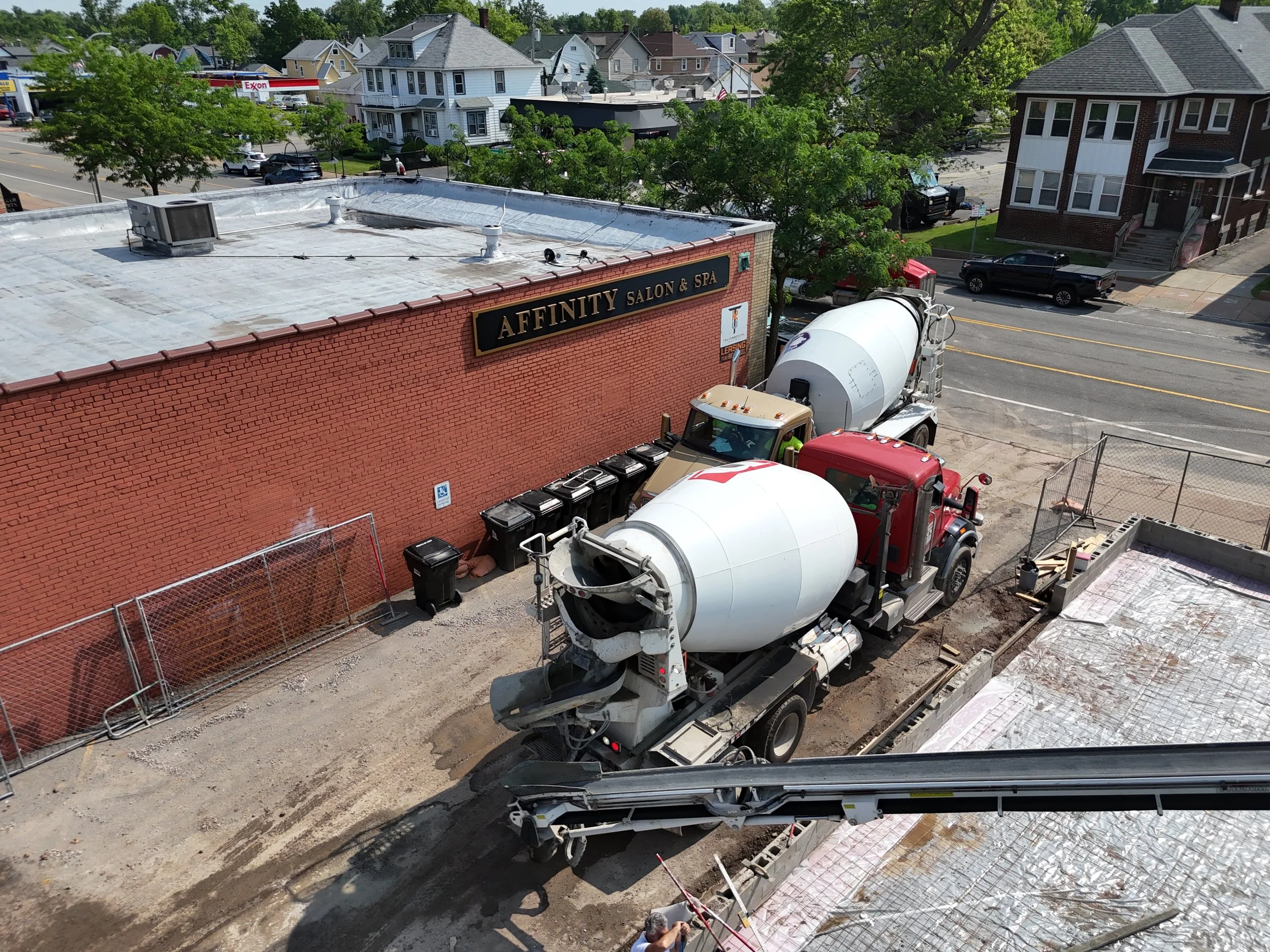 Construction site with cement mixer trucks pouring concrete in front of a brick building with a sign that reads 'Affinity Salon & Spa,' neighboring residential houses, and trees.
