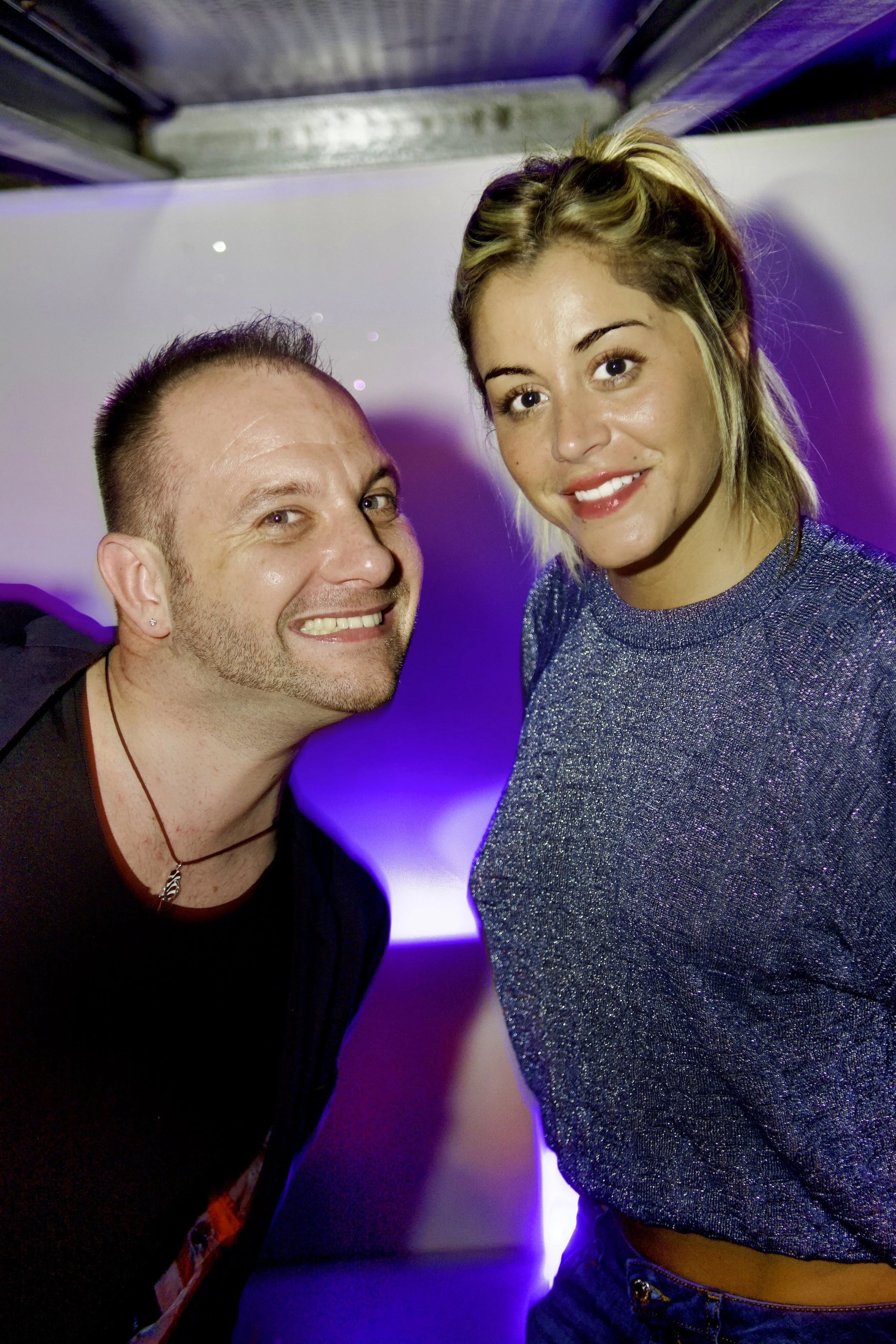 Deux personnes posent en soirée, l'une avec une coiffure courte et l'autre avec des cheveux blonds mi-longs, dans un environnement à éclairage violet.